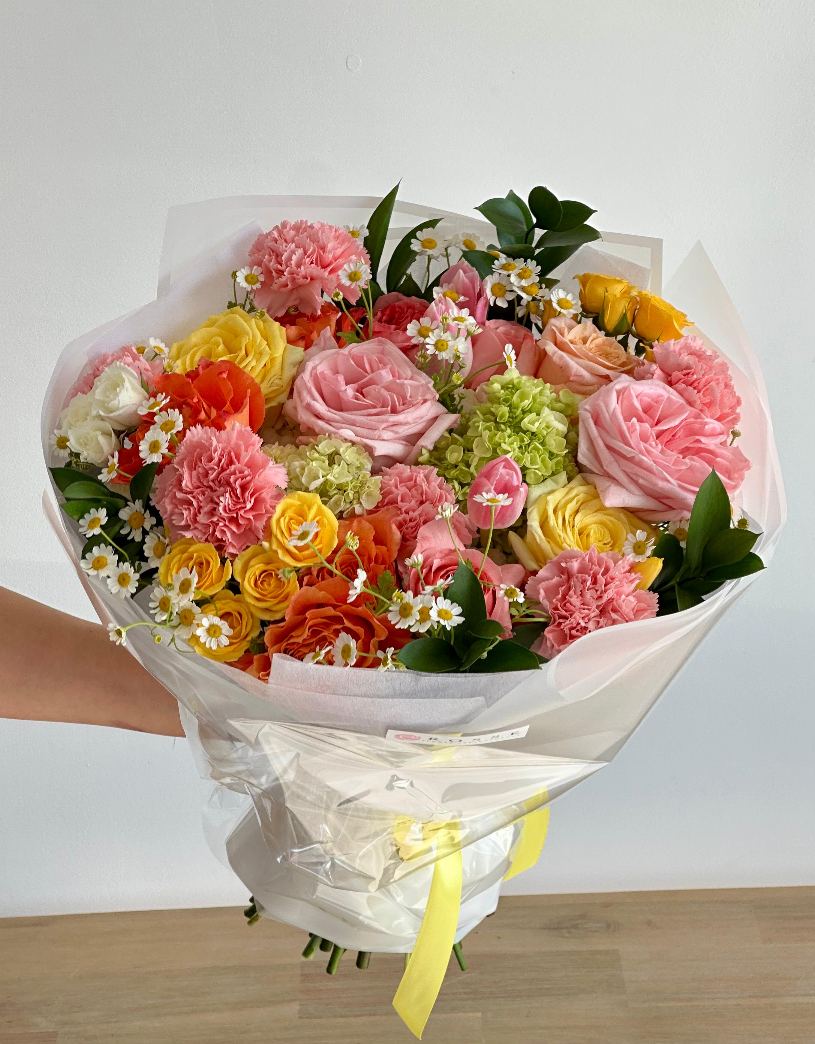 Handheld bouquet of pink, yellow, and orange flowers wrapped in clear paper