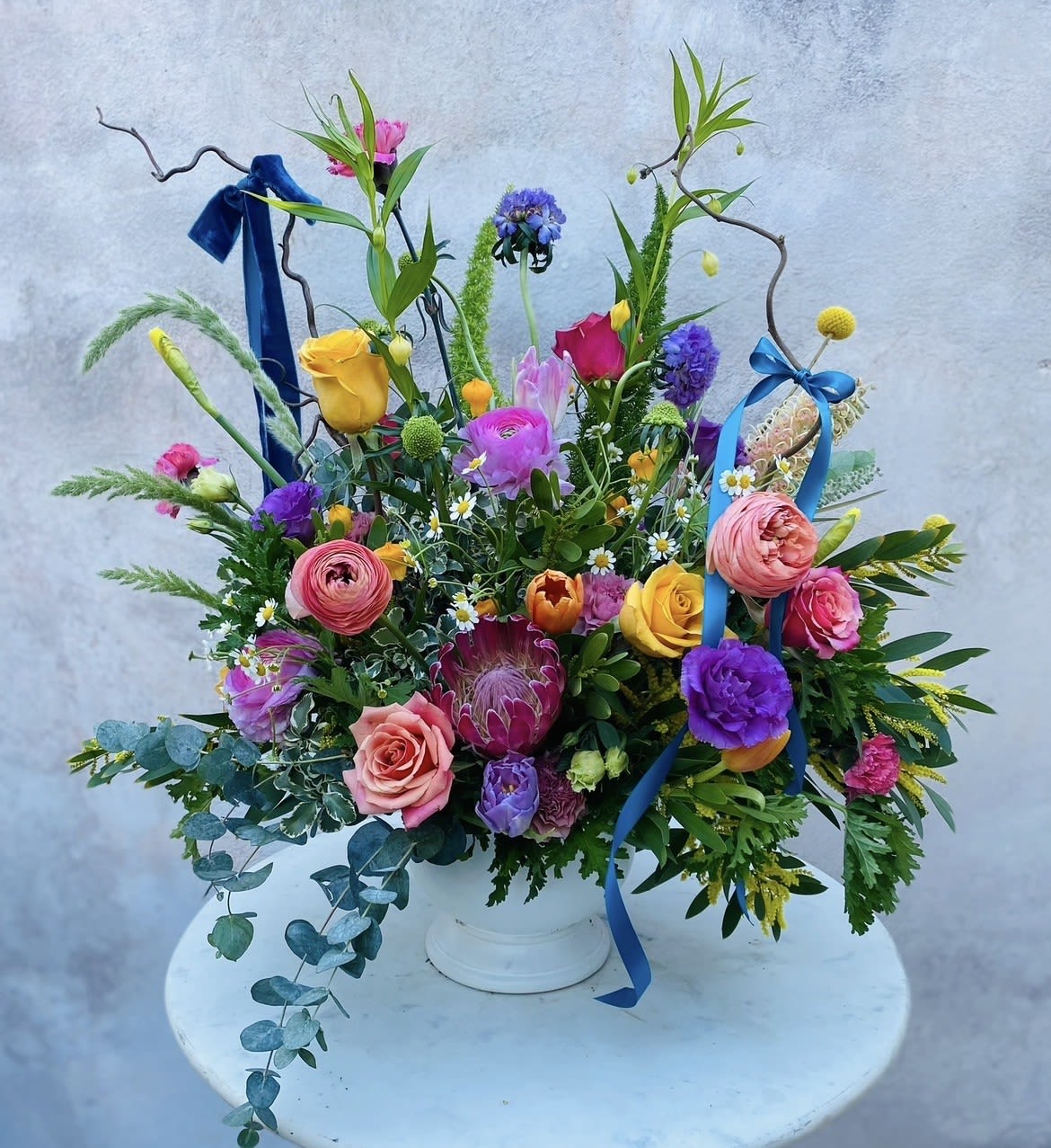 Maisie - A vibrant wildflower-inspired arrangement designed with fresh seasonal blooms and lush greenery. This bright, expressive design captures the natural beauty of a spring field, featuring a mix of airy texture, movement, and cheerful color.  Each arrangement is thoughtfully handcrafted using premium flowers selected for their seasonal character and unique form. The loose, garden-style composition creates a relaxed yet elevated look that feels fresh, natural, and full of life.  Flowers and greenery may vary due to seasonal availability, but that’s part of the charm! Our creative designers will substitute with the freshest high end product available- some grown in our very own garden!- and create something beautiful.  *Have a special request?* Let us know in the special instructions section during checkout.
