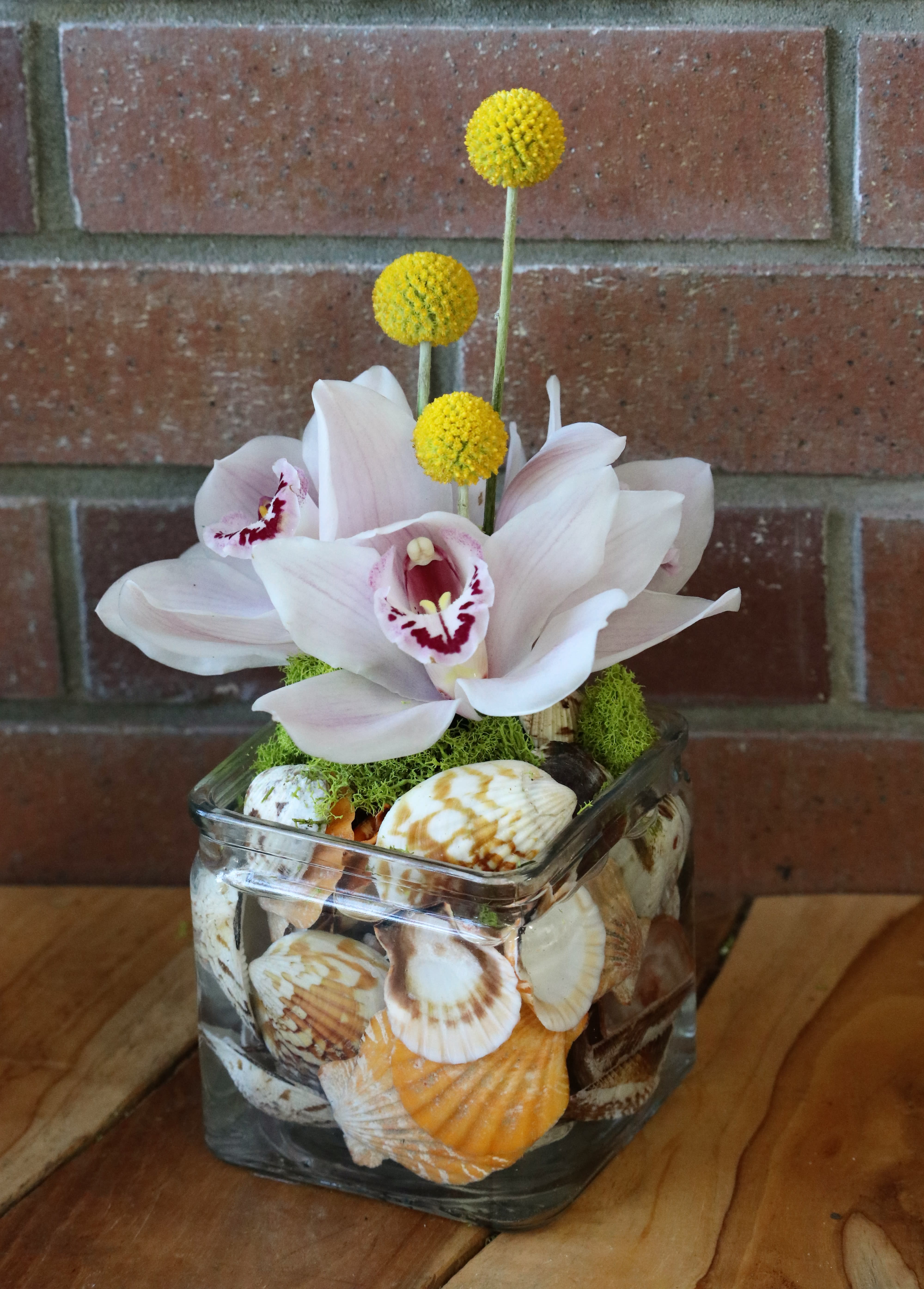Square glass vase filled with seashells topped with pale orchids and yellow ball flowers