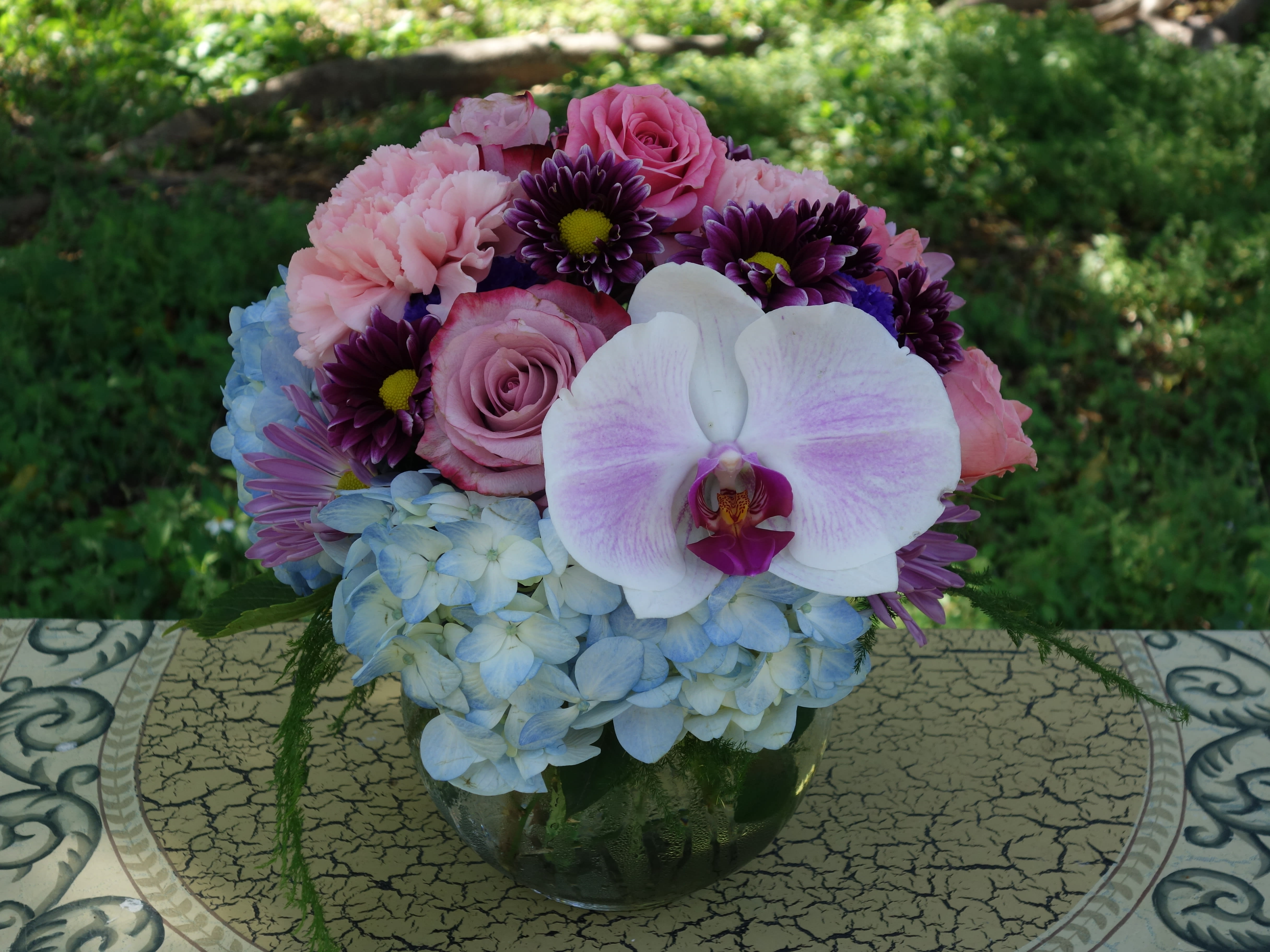 Round arrangement of pink roses and carnations with blue hydrangeas and a white orchid in a clear glass bowl