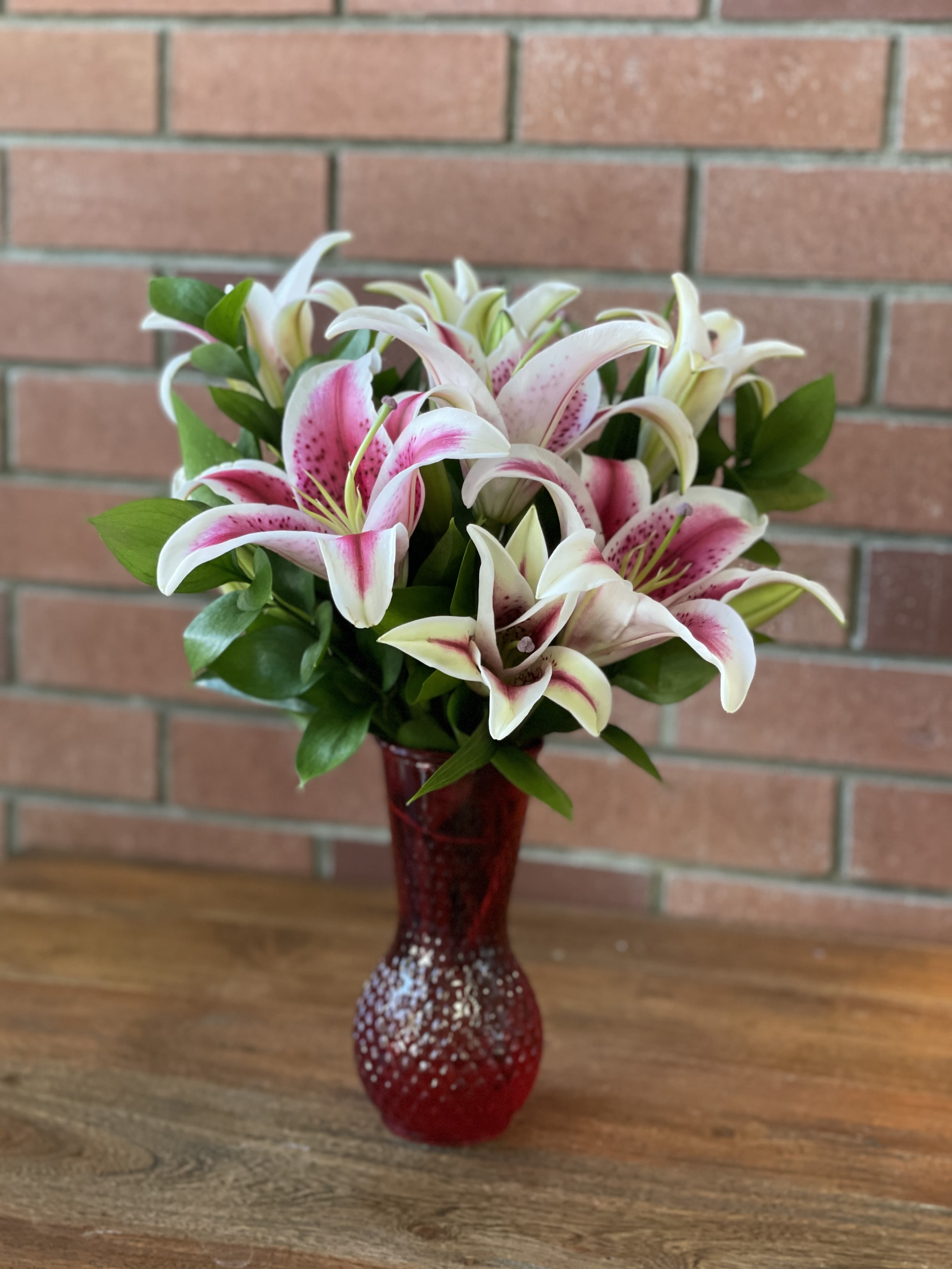 Pink and white lilies arranged in a red textured glass vase on a wooden surface.