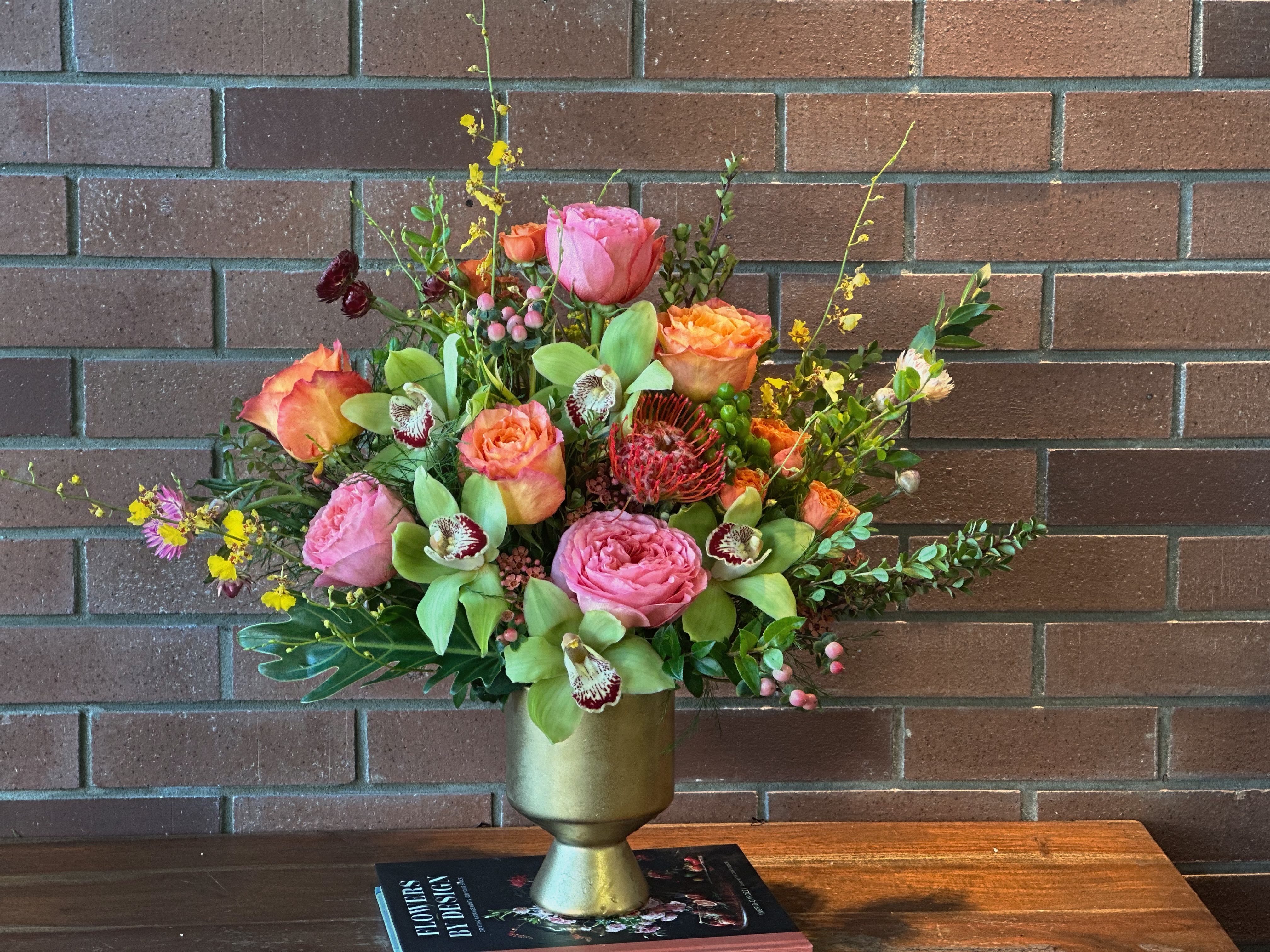 Sunset Lagoon  - A stunning symphony of seasonal blooms featuring lush pink garden roses, vibrant coral ranunculus, and exotic orchids, artfully arranged with textural elements including burgundy protea, delicate waxflower, and an array of fresh greenery. This sophisticated arrangement captures the essence of a romantic English garden with its rich color palette of coral, pink, and deep wine tones, complemented by golden accents and natural foliage. Presented in an elegant brass compote vessel, this design brings warmth and luxury to any space while celebrating the beauty of mixed seasonal botanicals.