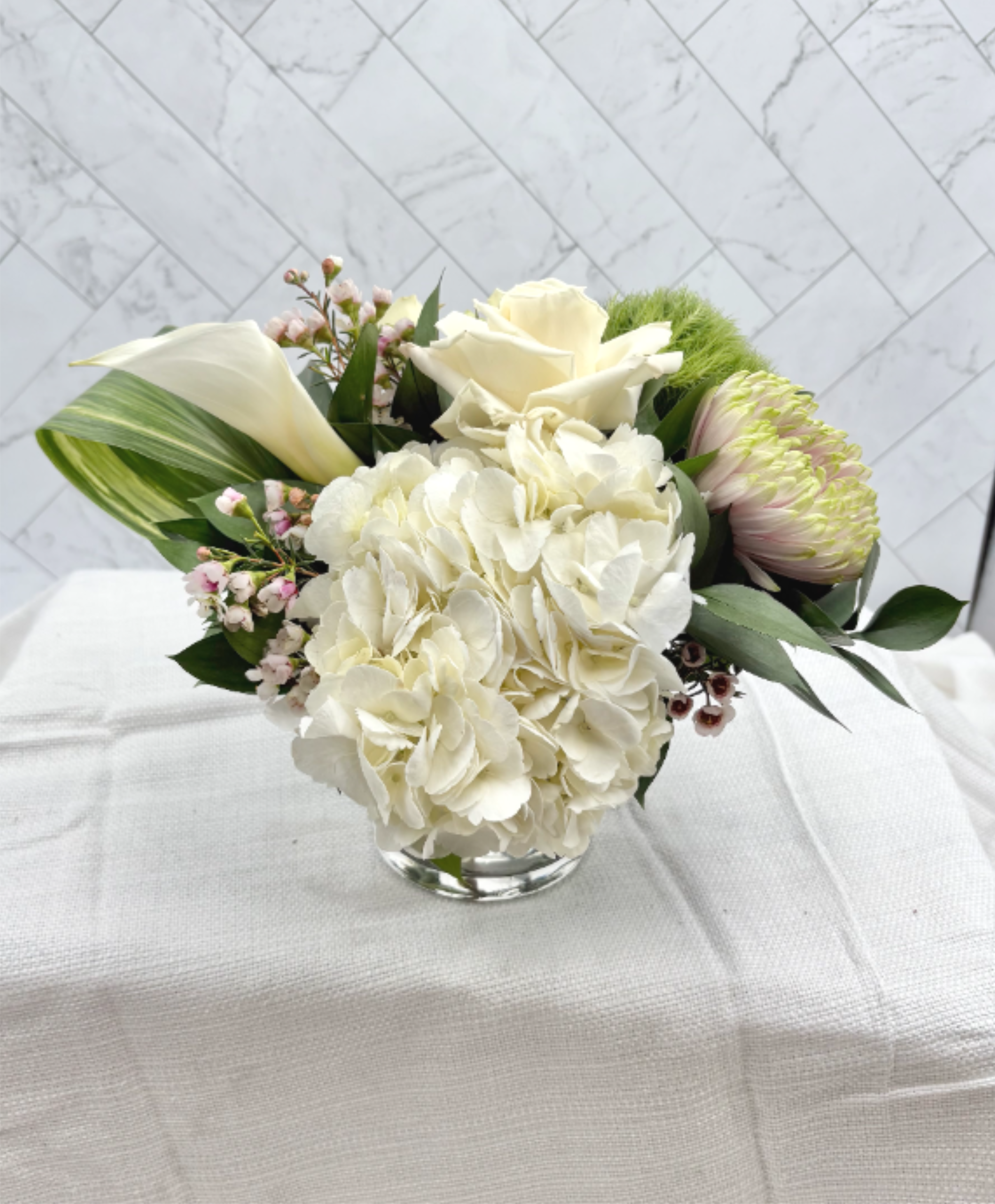 Low white floral arrangement with hydrangea, roses, and calla lilies in a clear glass vase