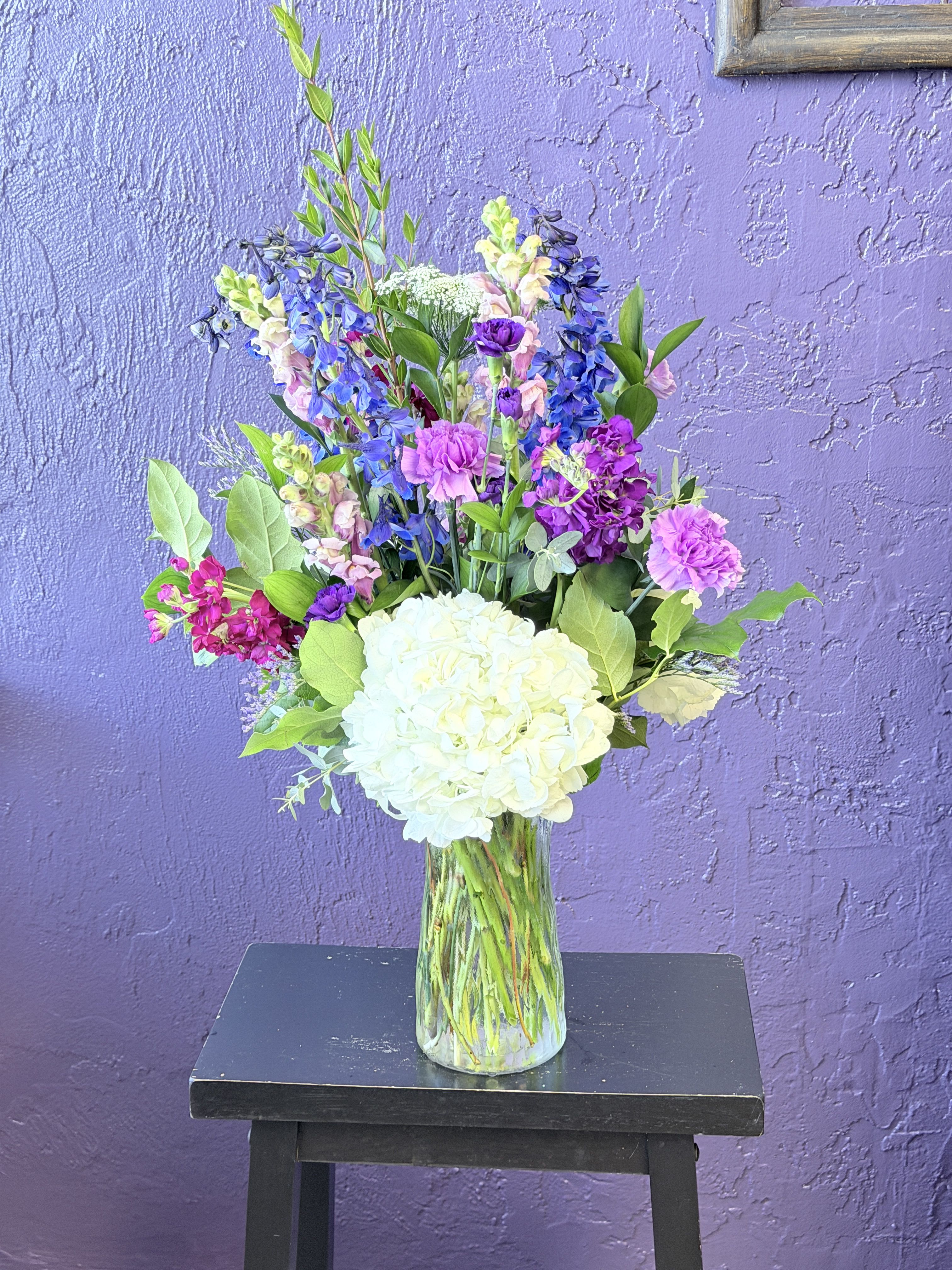 Heather Garden  - Tall spires of pink and lavender stock and snapdragons rise above soft clusters of blue, white, and pale-pink hydrangea to create a fresh, garden-style flower arrangement perfect for a Mother's Day bouquet, anniversary gift, or a birthday surprise. The vertical, airy silhouette feels upbeat and romantic-the kind of arrangement that makes the person you're thinking of feel seen and celebrated without being overdone.  This clear glass vase holds a layered mix of hydrangeas, stock, chrysanthemums, dianthus and other seasonal stems, with delicate branching filler adding texture and movement. The palette of pink, purple, white, and blue reads serene and cheerful; approximate dimensions are 12" diameter by 30" high, giving a substantial presence on a dining table or mantel.  Send this as a "just because" pick-me-up, a heartfelt thank-you, or a gentle Valentine's or Grandparent's Day surprise-it delivers garden-fresh charm and uplift. Vase care: keep water fresh, recut stems every few days, and place away from direct heat to prolong blooms.