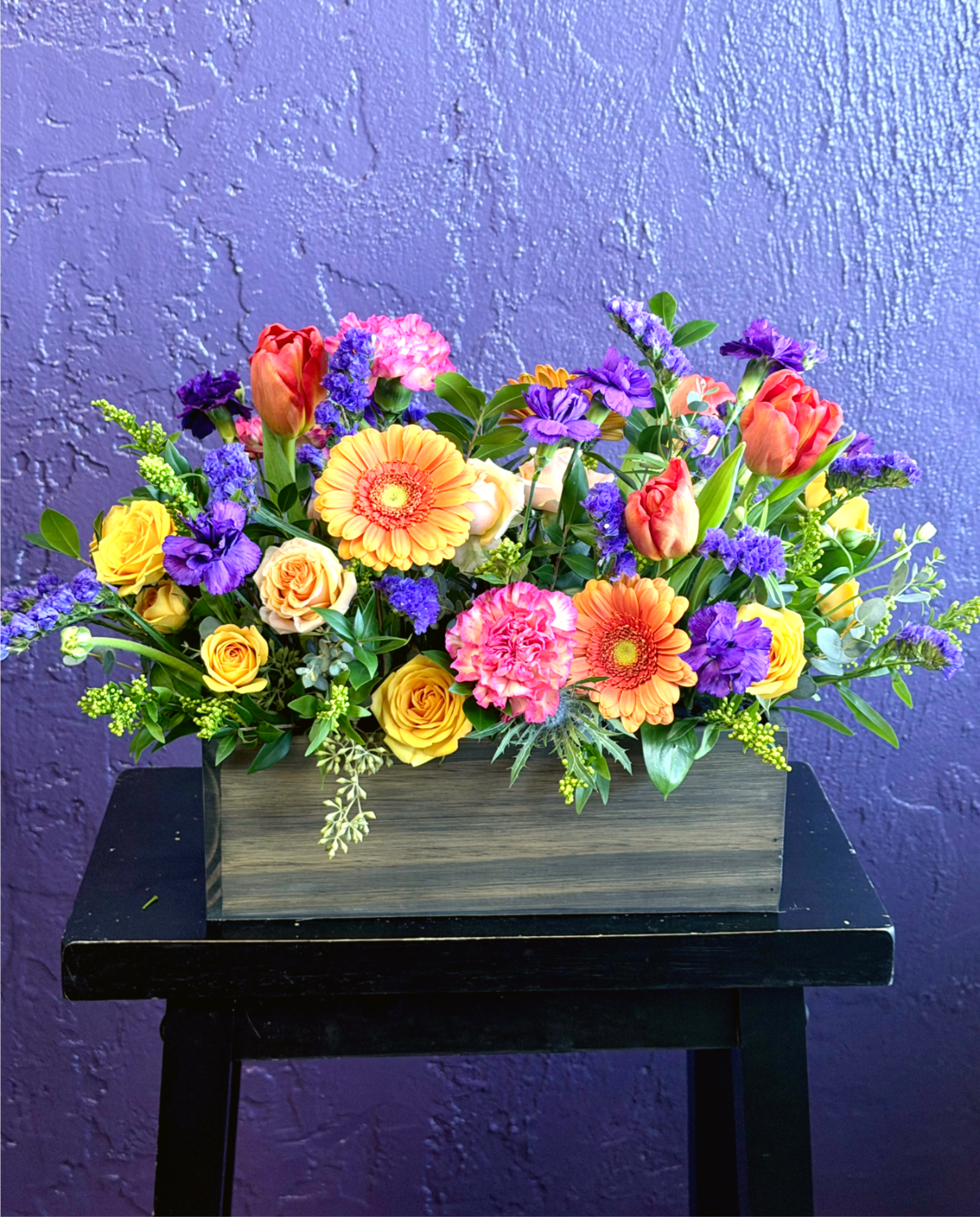 Groovy  - A vivid celebration of color, this mixed-color flower arrangement is made to make someone feel truly seen - perfect as a birthday bouquet, anniversary surprise, or a joyful Mother's Day gift. Bright pinks, purples, oranges, and sunny yellows combine to create an upbeat, festive look that also reads as thoughtful for graduations, new baby visits, thank-you gestures, or a spontaneous "just because" pick-me-up.  Choose between two presentations: a compact cluster in a green glass vase crowned with two tall bird-of-paradise stems for dramatic vertical impact, or a dense, low centerpiece in a weathered rectangular wooden box that spreads outward as a casual, artful display. Both include pink roses and other mixed blooms, orange gerbera daisies and textured tropical accents, coral tulips, pink carnations, purple stock and small daisy-like fillers for a layered, jewel-toned palette. Approximate size: 18" W x 10" H. Care: keep blooms in fresh water, trim stems every few days, and avoid direct heat to help the arrangement stay vibrant.