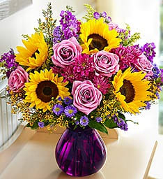 Floral Devotion - They’ve been there for you through it all. Let that special someone know how much they mean with our gorgeous bouquet of golden sunflowers and lovely lavender daisy poms &amp; roses. Hand-designed by our skilled florists inside a charming purple glass vase, this vibrant surprise is a touching expression of all the love you have in your heart.