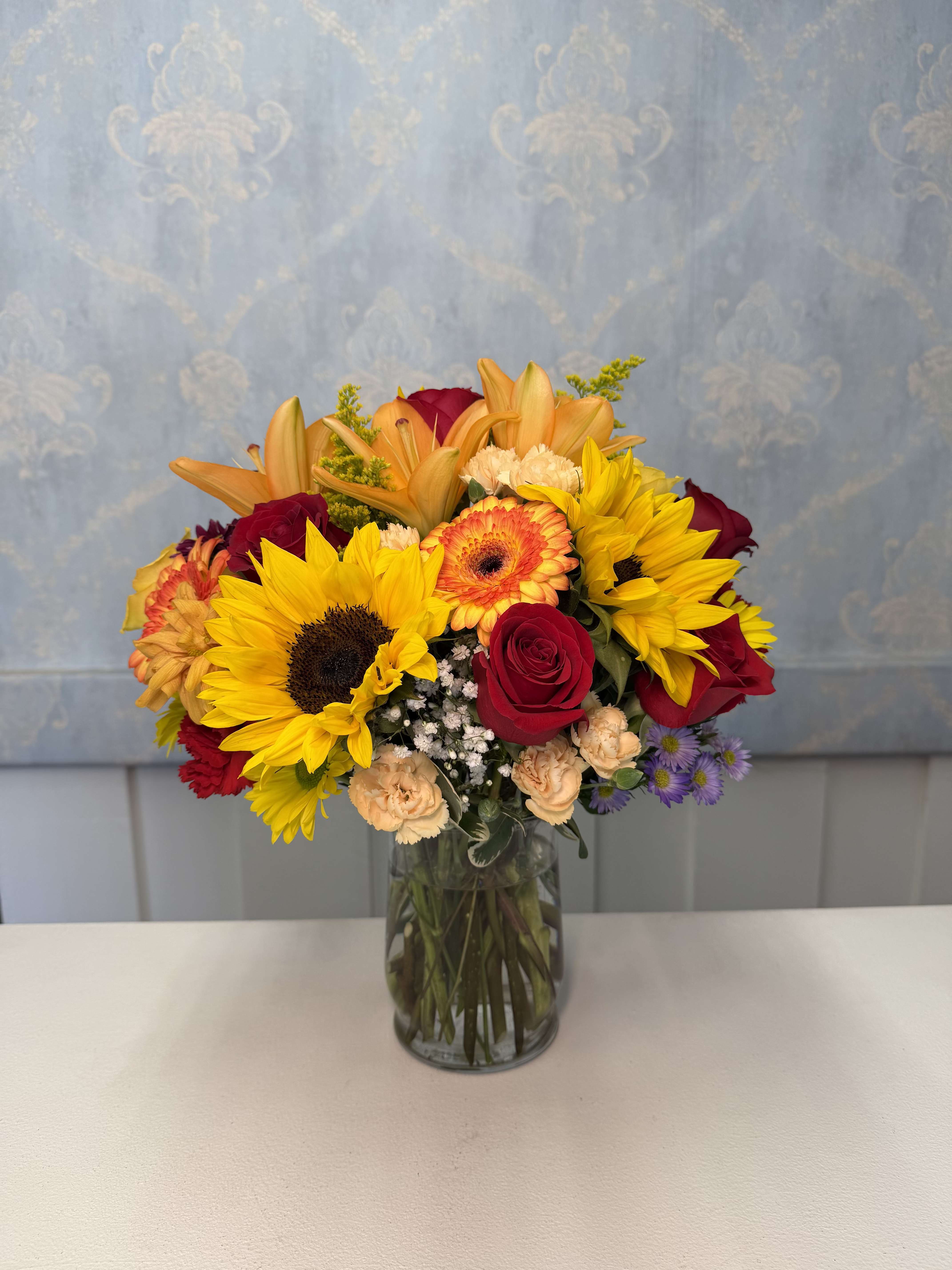 Golden Sunshine  - Brighten someone’s day with our stunning Golden Sunshine arrangement  A vibrant floral design featuring sunflowers, lilies, roses, gerberas, and a beautiful mix of fresh seasonal flowers arranged in an elegant glass vase.  This arrangement is full of happiness, color, and elegance — the perfect gift for birthdays, special occasions, or simply to make someone smile.    Flower colors and some varieties may vary depending on seasonal availability in our shop while always maintaining the overall style, size, and beauty of the arrangement.