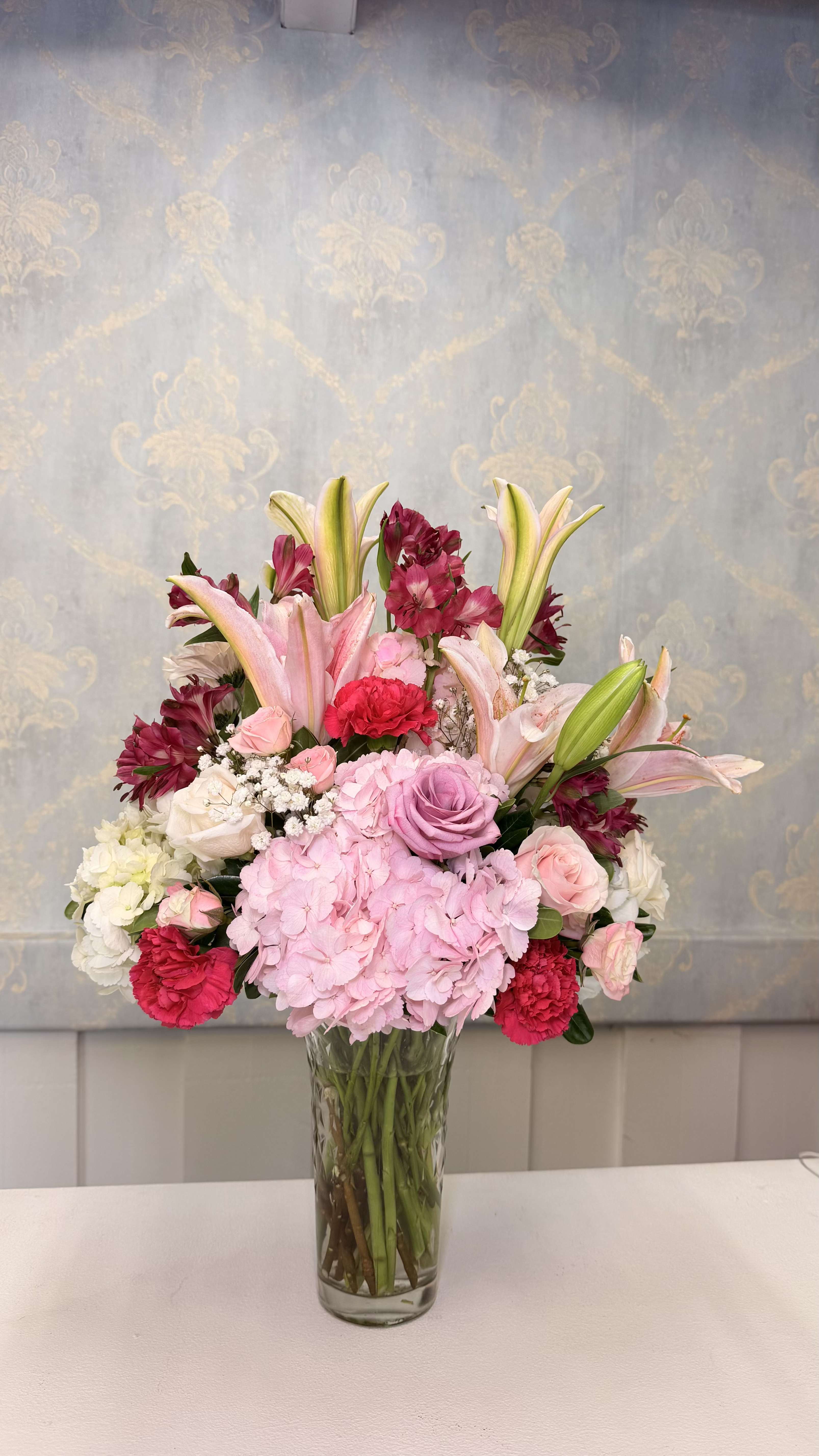 Blushing Elegance - Fall in love with the beauty and sophistication of our Blushing Elegance arrangement  This romantic floral design features stunning pink lilies, fresh hydrangeas, premium roses in different shades, delicate carnations, and seasonal flowers accented with lush greenery in an elegant glass vase.  Perfect for birthdays, Mother’s Day, anniversaries, or any special occasion, this arrangement is designed to make anyone feel loved and appreciated.    Flower varieties and colors may vary depending on fresh seasonal availability while always maintaining the overall style, size, and beauty of the arrangement.   A feminine, luxurious, and eye-catching arrangement that instantly brightens any space.