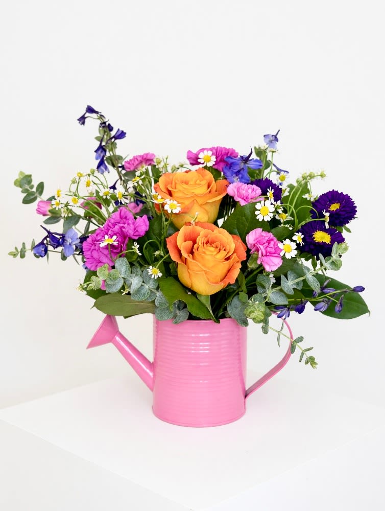Mom's Garden Bouquet - Bursting with color and personality, this cheerful garden‑style arrangement turns any day into a celebration. Lively orange roses, bold magenta and purple carnations, bright blue delphinium, and sunny white daisies gather in a charming pink watering can—creating a look that feels fresh, fun, and full of life. It’s the perfect blend of whimsy and warmth, like a little bouquet of sunshine delivered straight to the doorstep.