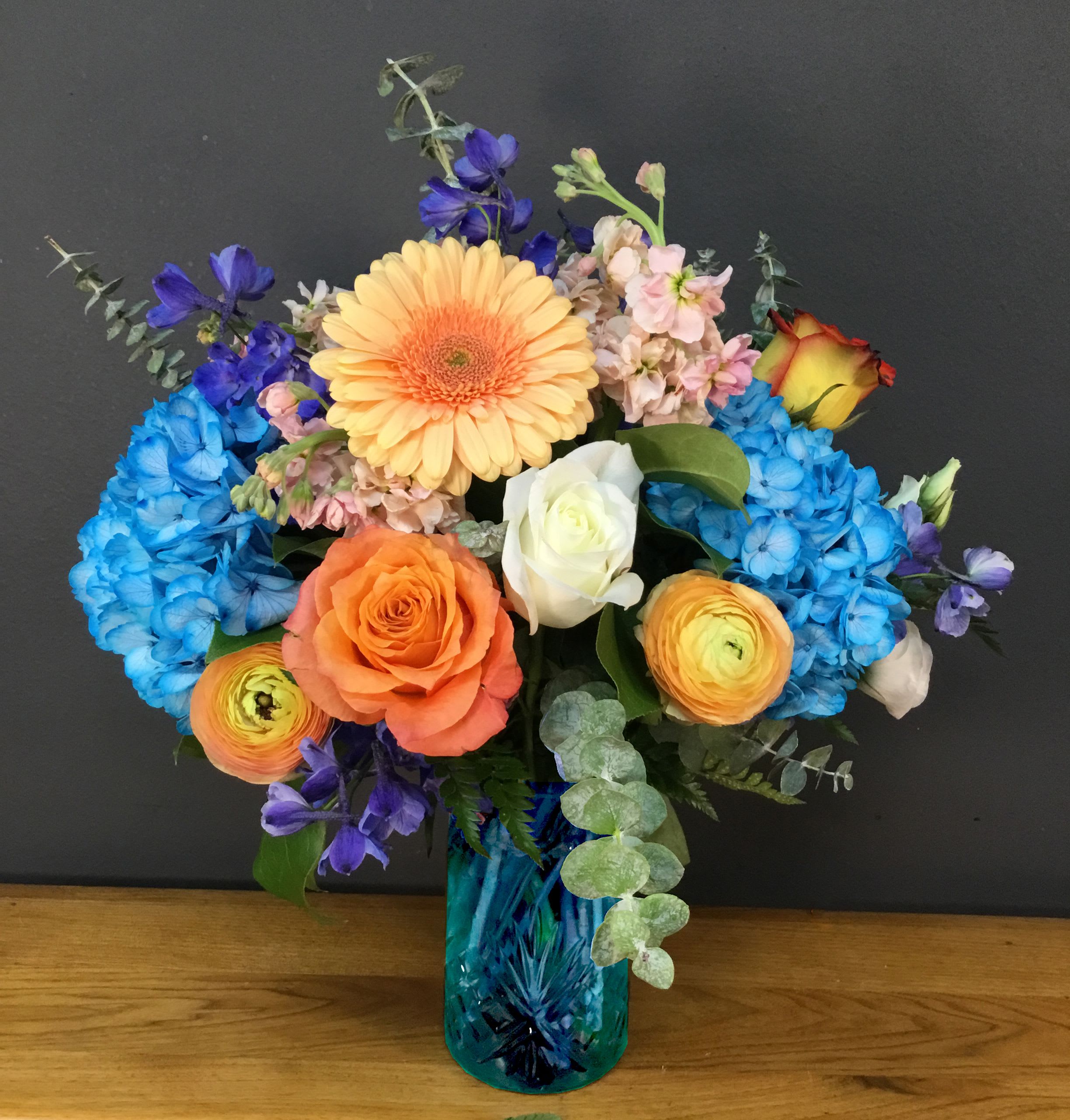 Blue Skies and Sunset - A bold, cheerful mixed bouquet that feels like a clear sky and a warm sunset in one vase-perfect as a birthday bouquet, congratulations arrangement, or an everyday celebratory gift. Blue Skies and Sunset combines bright blue hydrangeas and a prominent peach gerbera daisy at the center to create an immediate focal point, while orange, white, and yellow roses add sunny warmth that reads like an anniversary or special-occasion pick-me-up for the person you're thinking of.  This arrangement is gathered in a blue glass vase that complements the vivid blue hydrangeas and makes the pink and purple accent blooms pop. You'll also find ranunculus and other seasonal filler flowers layered throughout, with eucalyptus-like foliage softening the overall silhouette. The mix of textures-from the rounded hydrangea clusters to the daisy's cheerful face and the lush roses-creates a lively, layered look that fills a room with color.  Give this to someone who deserves to feel uplifted and celebrated-the bright palette says joy and thoughtfulness without being overly formal. Arrives ready to display in the blue glass vase pictured; trim stems and refresh water every few days to keep blooms looking their best.