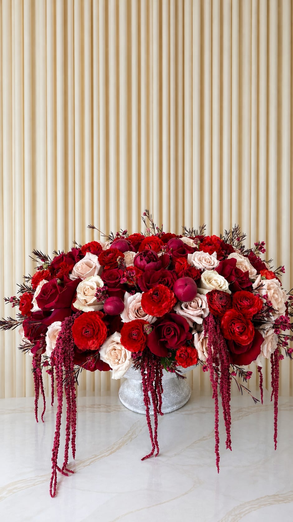 Cascading Reds - A lush cascade of saturated reds, blush pinks, and creamy whites creates a romantic flower arrangement perfect for an anniversary bouquet, Mother's Day gift, or any moment you want someone to feel deeply celebrated. This large, low, densely packed design places red roses and blush roses at the center, joined by peonies and delicate ranunculus, while deep burgundy accents and draping amaranthus-like strands give it a dramatic, cascading silhouette that reads as both passionate and refined.  Dense, closely grouped blooms sit in a concrete vase so the arrangement is sized to impress yet made for tabletop display on a dining table, entryway, or bedside. The mix of saturated reds and softer pale tones creates striking contrast and elegant balance; the trailing amaranthus adds movement and the ranunculus and peonies bring soft texture and lush volume. Arrives ready to brighten the room.  Ideal for anniversaries, romantic celebrations, and Mother's Day, this bouquet sends a message of devotion and thoughtful attention-the kind of gift that makes the person you're thinking of feel truly seen. For best enjoyment, keep the vase filled with fresh water and trim stems every few days to maintain the dense, lush look.