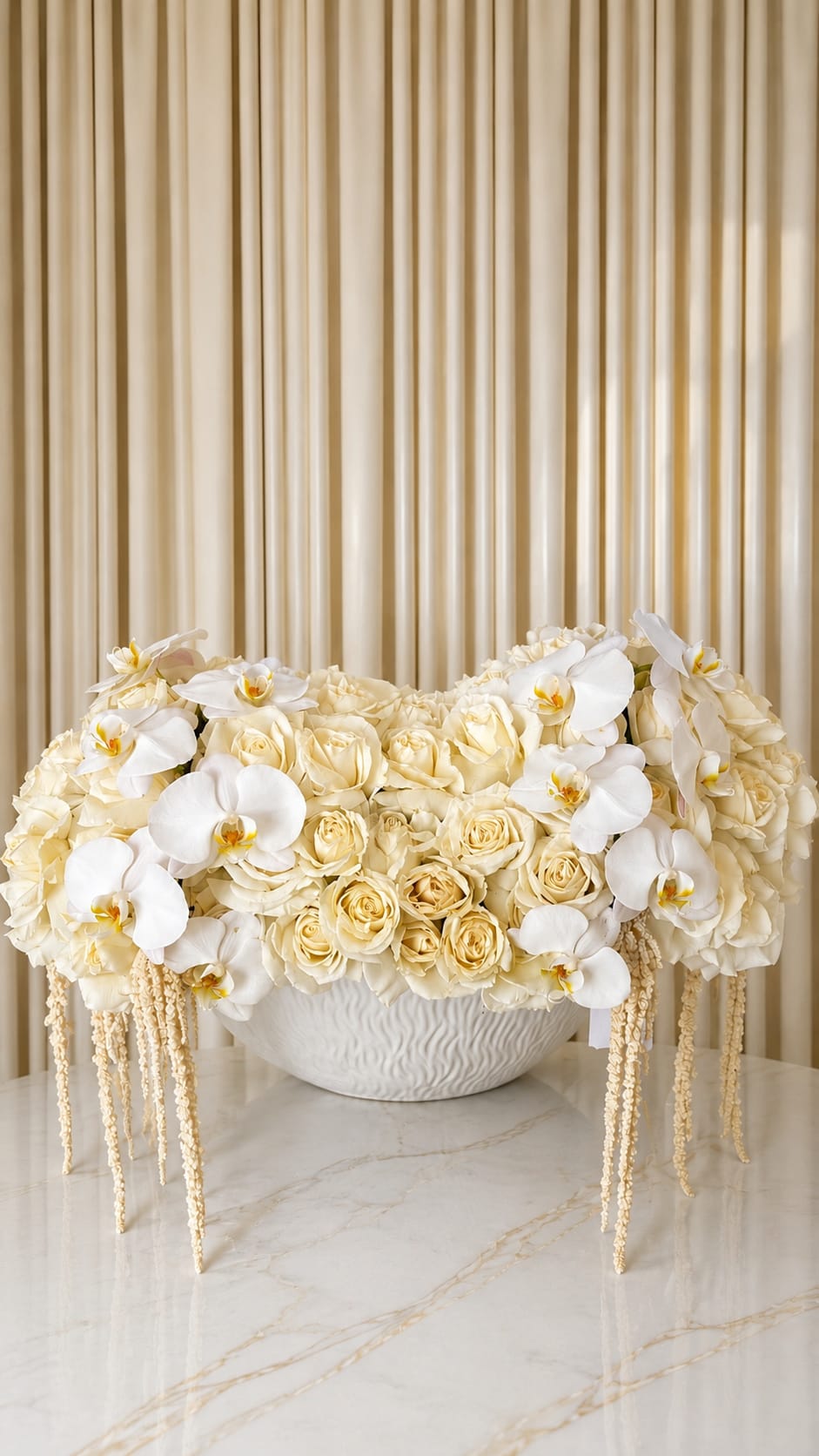 Whisper Of Whites - A timeless expression of elegance, this luxurious arrangement blends cascading white orchids with layers of premium ivory roses in a sculptural textured vessel. Designed with soft movement and refined balance, each bloom is carefully placed to create a serene, cloud-like composition that feels both modern and romantic. Delicate hanging amaranthus adds graceful dimension, while the creamy monochromatic palette brings a sense of warmth, sophistication, and effortless luxury. Perfect as a statement centerpiece for intimate gatherings, grand celebrations, or elevated home décor, this arrangement embodies understated beauty with an artful, couture-inspired touch.