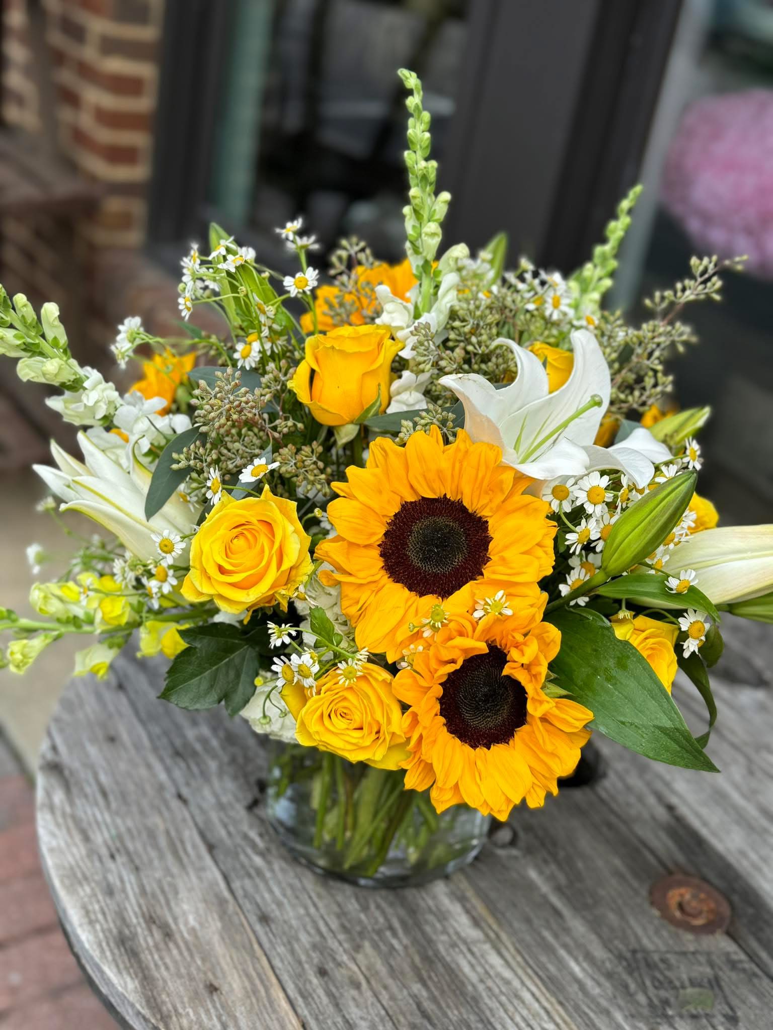 Sunshine Vase - Bright, sun-kissed blooms take center stage in this cheerful flower arrangement-perfect as a birthday bouquet, a bright thank-you gift, or a spring celebration centerpiece. The Sunshine Vase sends an uplifting message to the person you're thinking of: warmth, joy, and a little sunshine to brighten their day.  A full, layered composition of yellow roses and bold sunflowers is softened by white lilies and small daisy-like filler flowers, with green accents woven throughout for a fresh, garden-style look. Arranged loosely in a clear glass vase, the mixed textures-velvety rose petals, sturdy sunflower heads, elegant lily blooms, and spiky snapdragons-feel natural and hand-gathered, about the size of a generous table bouquet suited for display on a mantel or dining table.  Give this arrangement to someone who could use a sunny lift-the kind of gift that makes them feel genuinely seen and celebrated. Vase-ready on arrival with simple care: keep water fresh, trim stems every few days, and place out of direct heat to extend bloom life.