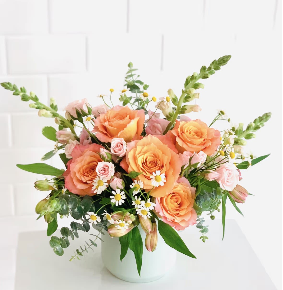 Summer Sunset - Peach and pink roses take center stage in this Summer Sunset flower arrangement, a birthday bouquet and spring celebration choice that reads cheerful and romantic at first glance. The rounded, airy design feels freshly gathered-perfect for saying "happy birthday," "thinking of you," or celebrating warm-weather moments-so the person you're sending it to will feel seen and genuinely celebrated.  Soft white daisy-like blooms, pale pink alstroemeria accents, and loose green foliage create a garden-like texture that spills naturally from a simple white ceramic vase. The palette of pink, peach, white, and green keeps the mood light and versatile, while the varied bloom shapes-full roses, delicate daisies, and trumpet-like alstroemeria-give the arrangement a lively, tactile depth.  Designed for everyday gifting as well as special springtime occasions, Summer Sunset arrives ready to display in its compact white ceramic vase. For best longevity, place in cool water away from direct sunlight and trim stems slightly before arranging; this keeps the bouquet looking freshly gathered for days.
