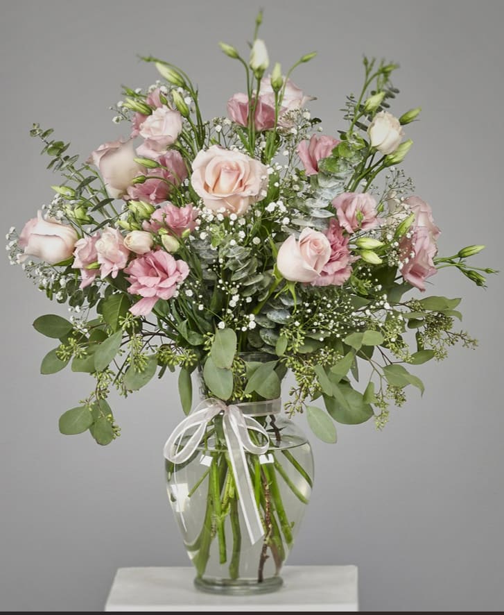 Tall arrangement of pale pink roses and lisianthus with baby's breath in a clear glass vase tied with a ribbon.