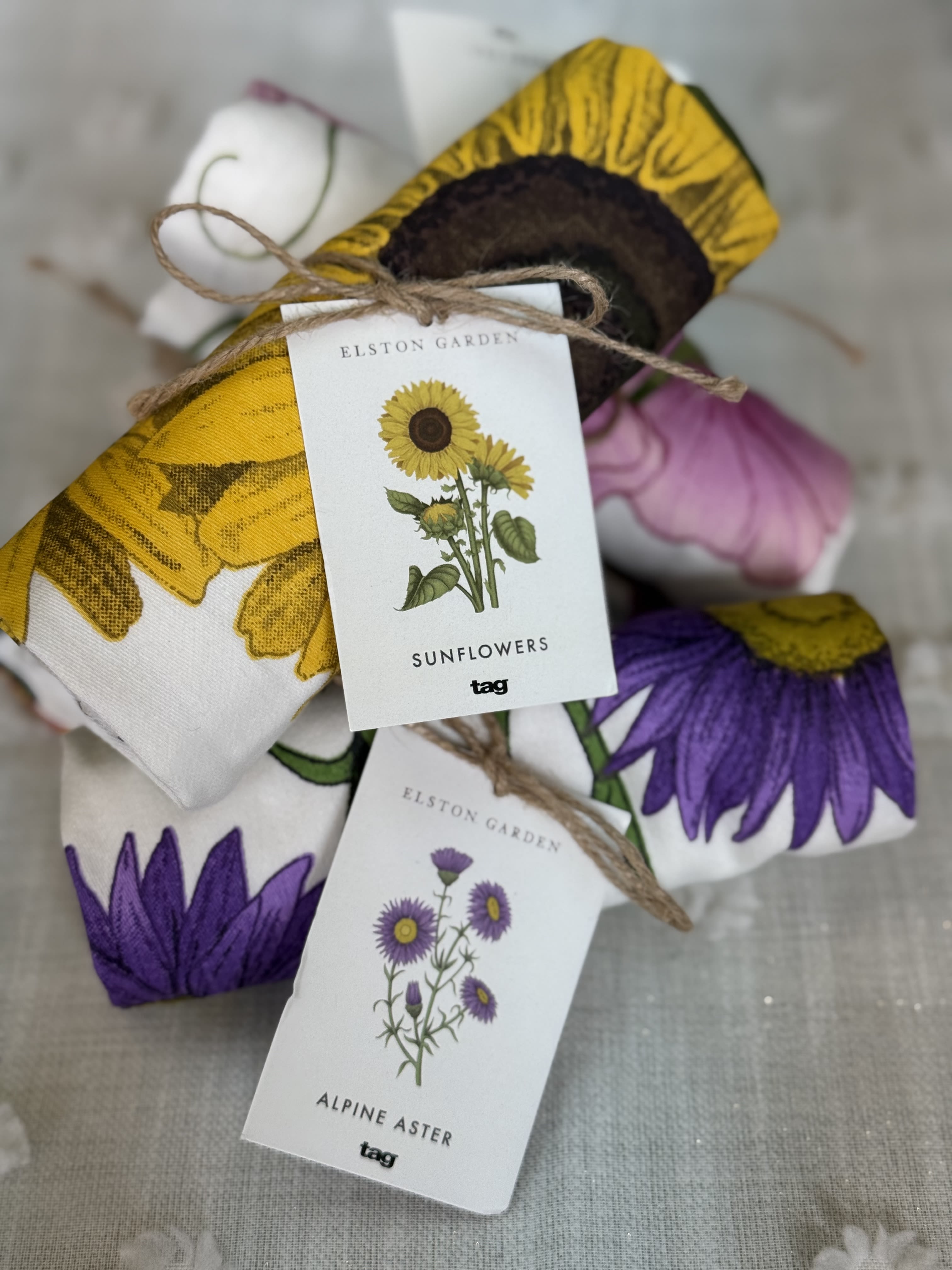 Kitchen Towels - Bold sunflowers and delicate purple asters print across soft kitchen towels to create an instantly cheerful housewarming or birthday gift that also suits Mother's Day, spring and summer occasions, and everyday thinking-of-you moments. The bright yellow, violet, white, and touches of black read like a lively garden on fabric, so the person you're gifting will feel noticed and uplifted by the sunny, handpicked look.  Each set includes folded fabric items bundled with natural twine and finished with branded paper tags for a boutique, handmade touch. The cotton-like texture shows clear sunflower and aster illustrations; the compact folds make them perfect for display on an apron hook, countertop, or in a gift basket alongside holiday or hostess presents. Packaged ready to give, this casual gift is equally at home for housewarming, graduation, retirement, Thanksgiving prep, or a cheerful get-well surprise.  Ideal for people who love bright, seasonal accents, these towels are easy to care for-wash gently to preserve the printed motifs-and arrive tied and labeled for immediate gifting. Whether you're celebrating an anniversary, saying thank you, or simply bringing a little sunshine to someone's kitchen, this bundle feels thoughtful and ready to use.