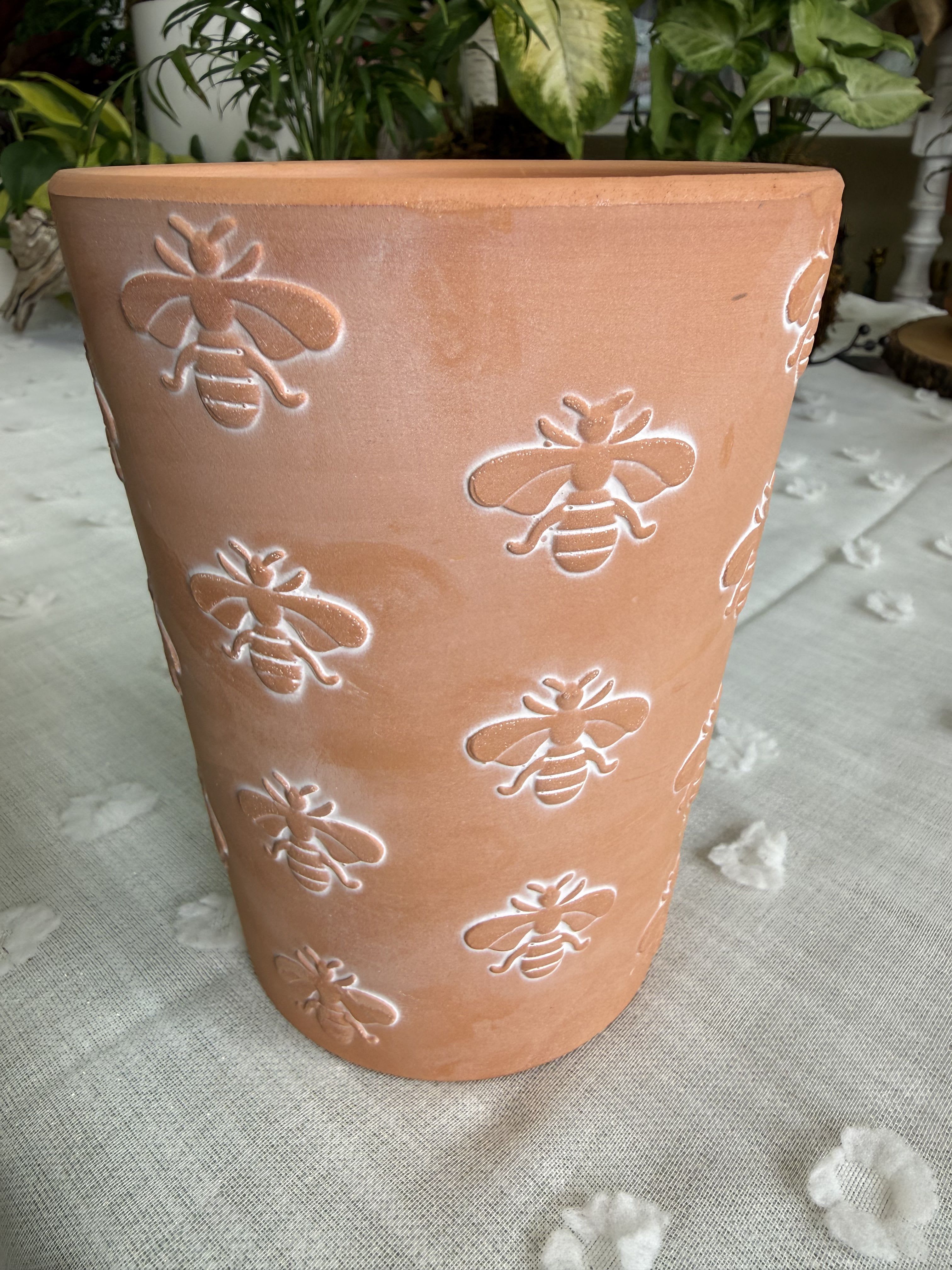 Bee Clay Pot for Plants - Sun-warm terracotta and cheerful bees make this tapered planter an instantly inviting choice for a housewarming gift, Mother's Day surprise, or a just-because pick-me-up. The flower arrangement-ready terracotta planter is embossed with repeated bee motifs highlighted in white, giving it a playful garden feel that suits autumn porch displays, spring patio plantings, or a cozy indoor accent for Thanksgiving, Christmas, or everyday décor.  The medium-to-large tapered planter has a matte, rustic finish that reads handmade and earthy-its orange-brown surface brings out greenery and orange or white blooms beautifully. Use it to pot seasonal mums, a mixed green and flowering arrangement, or a statement succulent grouping; the white-highlighted bees add contrast that elevates both bold and soft palettes. Its size makes it right for graduations, anniversaries, birthdays, get-well wishes, and retirement celebrations when you want the recipient to feel genuinely seen.  Practical and pretty, this planter works for holiday gifting, sympathy tables, or garden-loving hosts-perfect for Memorial Day, Father's Day, Labor Day, Halloween, or any special moment from prom to new baby. Pair with a favorite plant or let us fill it for delivery; simple care for planted specimens keeps the display thriving so your recipient enjoys the thoughtful gesture long after the occasion.