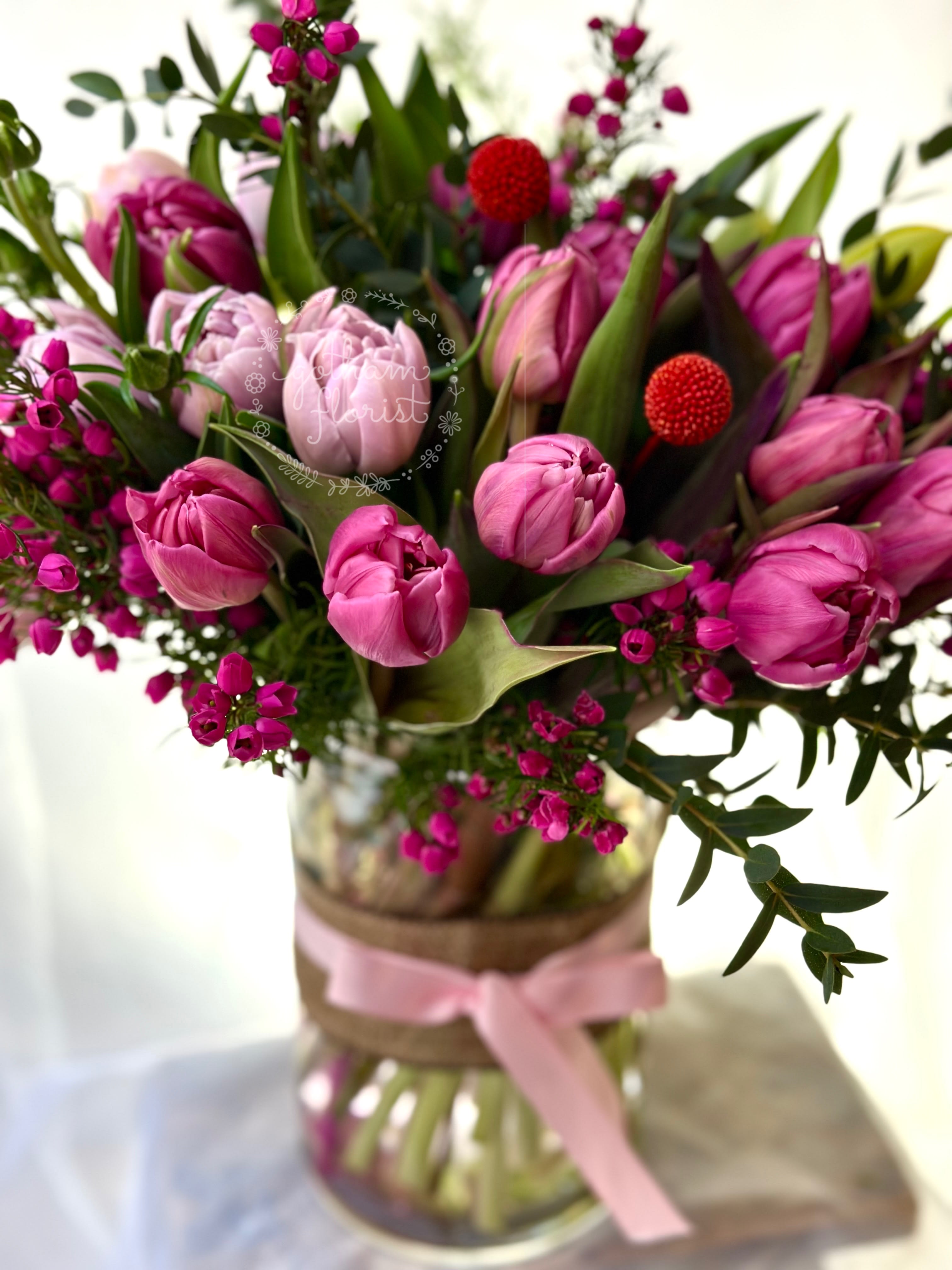 Pink tulips and magenta flowers arranged in a glass vase with a pink ribbon