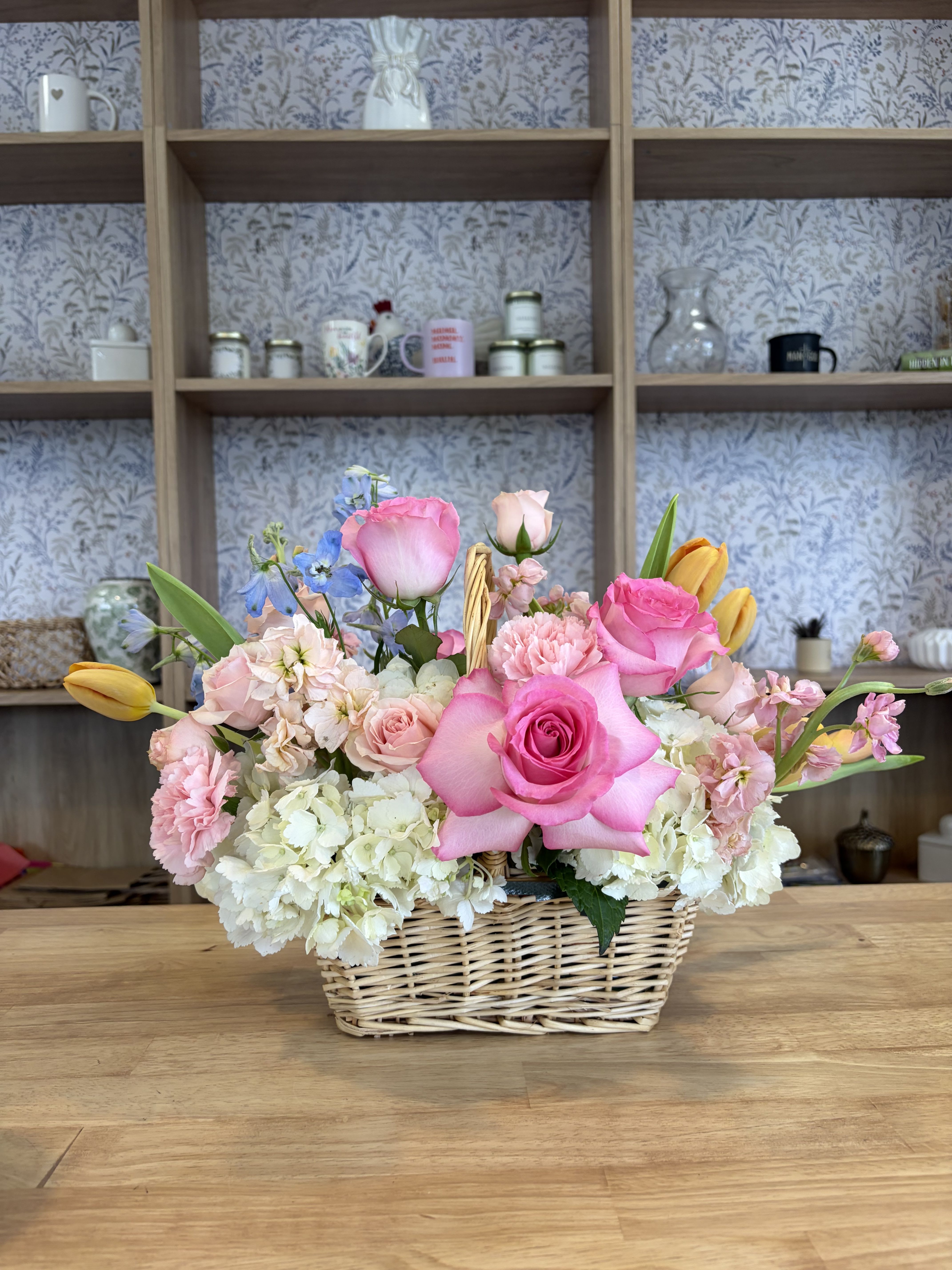 Blooming Basket - A garden-fresh mix of pastel roses and spring blooms makes this flower arrangement an effortless way to celebrate someone special. The Blooming Basket is a flower arrangement ideal for birthdays, Mother's Day, or "just because" moments-she'll feel genuinely seen when greeted by the soft pink roses and cheerful pops of yellow tulips framed in a low wicker basket.  This full, rounded composition pairs pink roses with fluffy white hydrangeas, pale carnations, blue delphinium, tulips, and stems of stock for scent and texture. The palette of pink, white, yellow, and blue creates a fresh, romantic look that reads as both casual and thoughtfully arranged; brighter pink focal roses draw the eye while the delphinium adds vertical contrast. The wicker basket gives a homey, gift-ready presentation and keeps the overall silhouette compact and table-friendly.  Perfect for brightening a kitchen table, sending as a thoughtful congratulations, or surprising a friend who loves cottage-garden style, this arrangement arrives ready to display. For best life, keep the arrangement in a cool spot away from direct sun and top up the water as needed-its low basket shape makes it easy to place anywhere the recipient will see it.