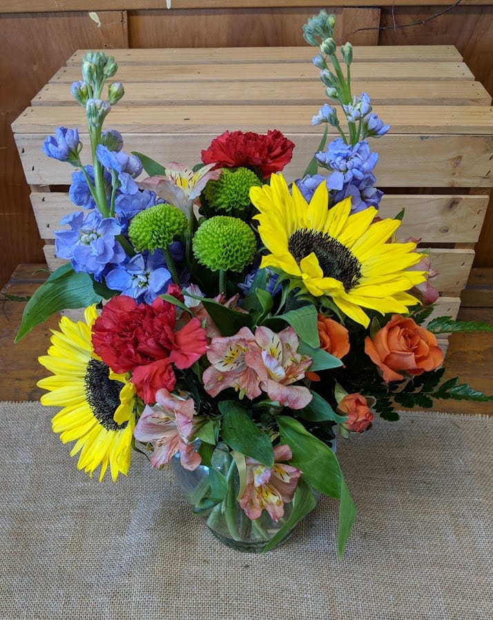 Mixed bouquet with sunflowers, roses, carnations, and blue flowers in a glass vase