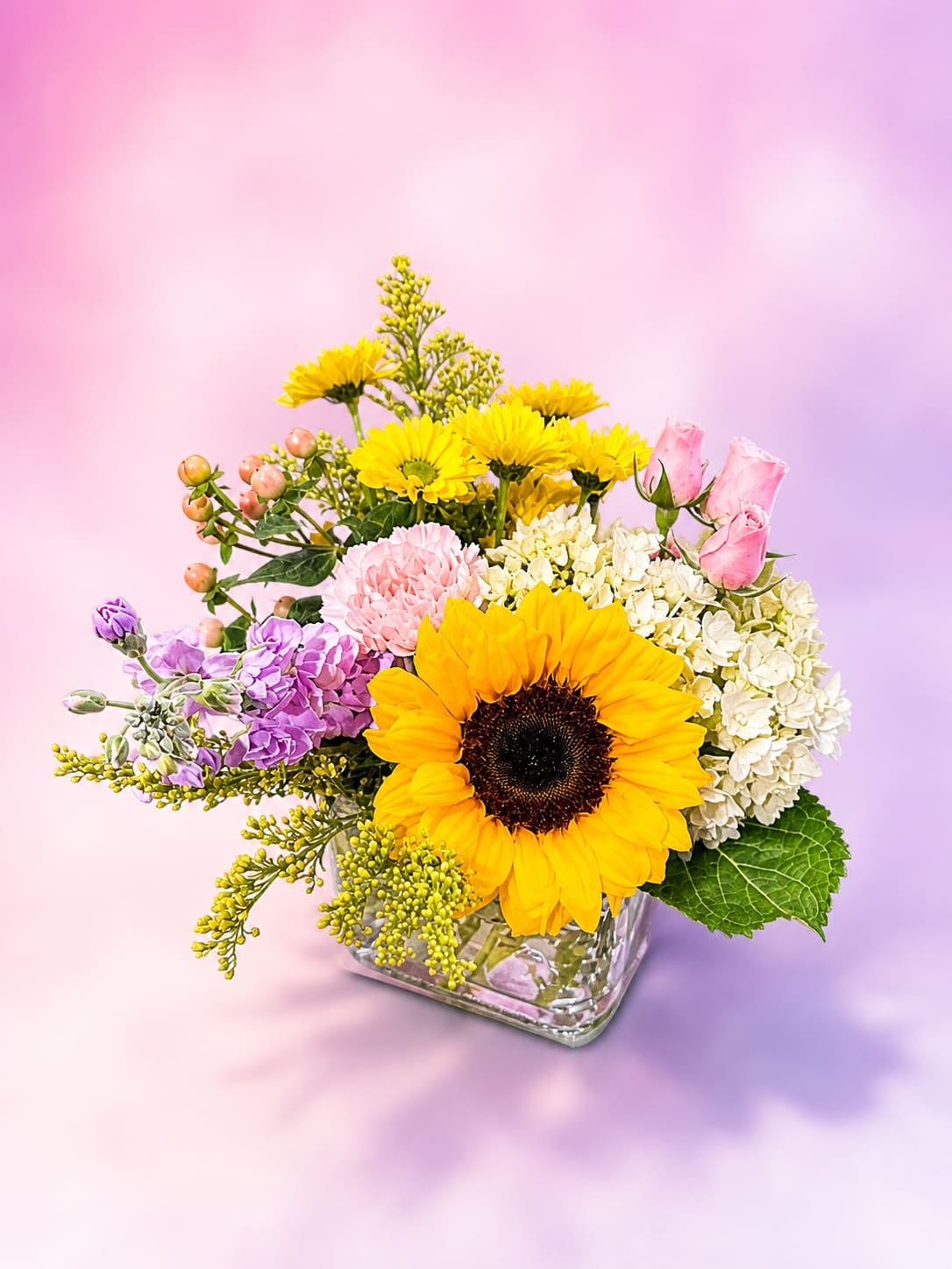 Sunshine and Sweet Stems - Bright, cheerful, and full of charm, Sunshine &amp; Sweet Stems features a radiant sunflower surrounded by soft pastel blooms and fresh seasonal textures. Accented with delicate roses, hydrangea, stock, carnations, and sunny yellow daisies, this arrangement brings together warm golden tones with soft pinks and lavender for a perfectly balanced garden-inspired design. Arranged in a modern clear glass cube vase, it’s a happy and uplifting piece perfect for birthdays, Mother’s Day, get well wishes, or simply bringing a little sunshine indoors. Arrangement is available in clear or red vase depending on availability.