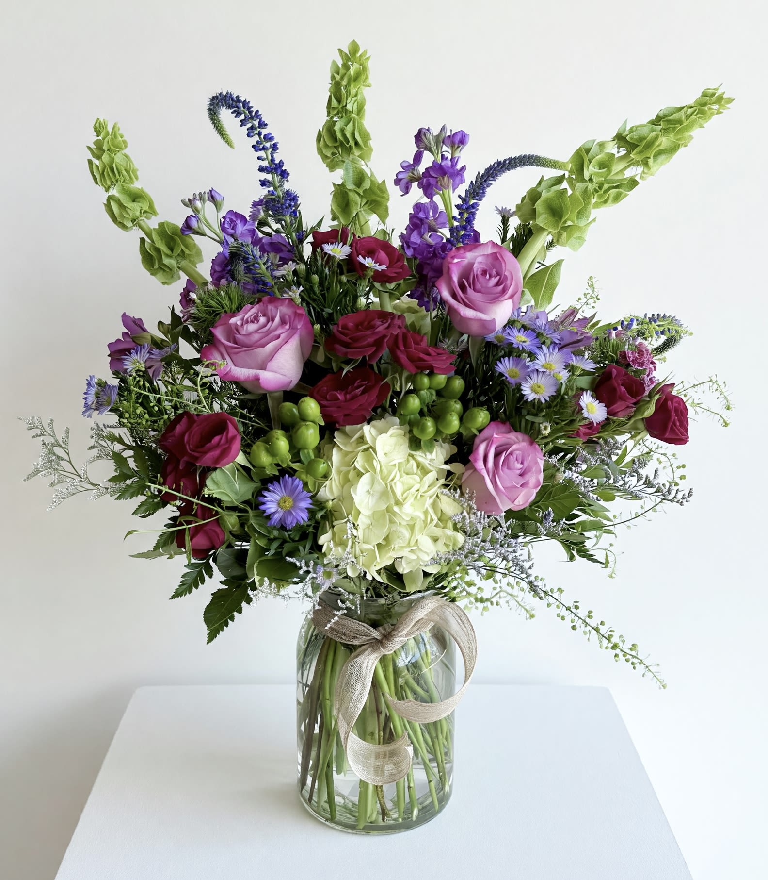 The Luka  - A clear glass vase filled with jewel tones of purple, lavender, red and green. A seasonal mix of flowers in these colors and will include stock, roses, green hydrangea, berries and other seasonal varieties. 