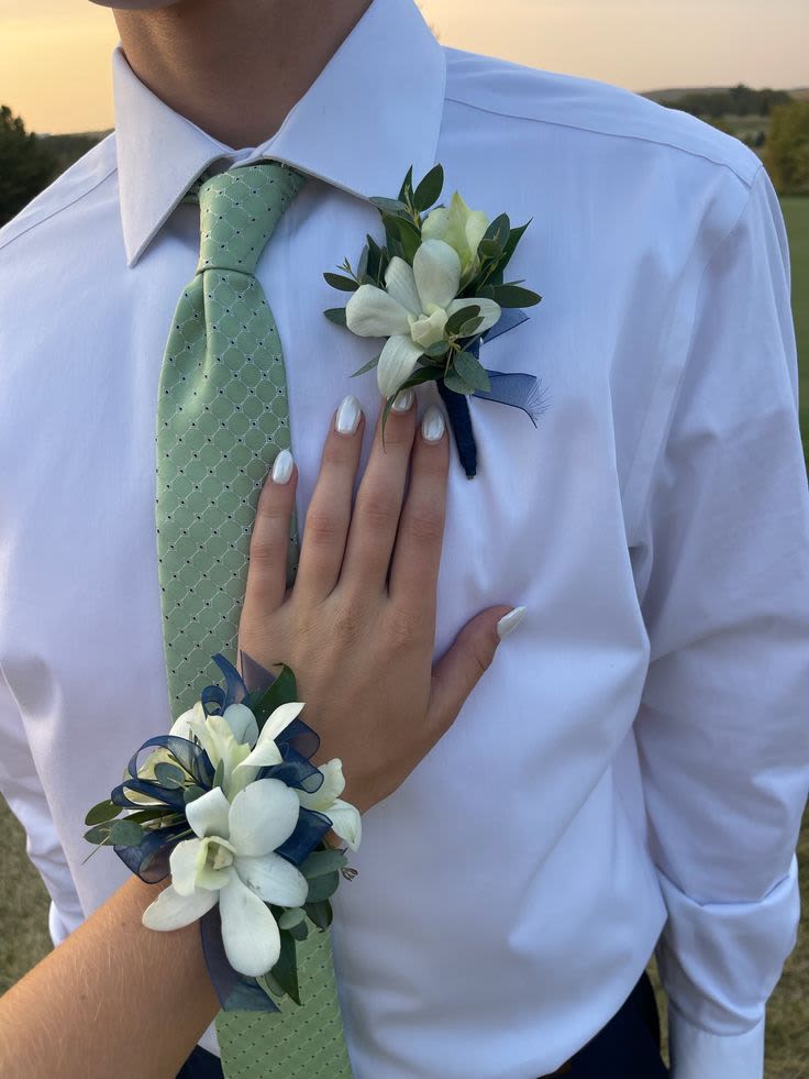 Green Orchid Corsage &amp; Boutonniere Set - This matching corsage and boutonniere set features fresh white orchids, soft greenery, and elegant sheer ribbon accents for a modern and sophisticated prom look.  Designed with fresh flowers and carefully coordinated details, this set brings a clean and timeless touch to your special night.  Includes: • Wrist corsage • Matching boutonniere • Fresh flowers • Ribbon detailing  If you would like a different ribbon color to match your outfit or prom theme, please leave your preferred color in the notes section at checkout.  