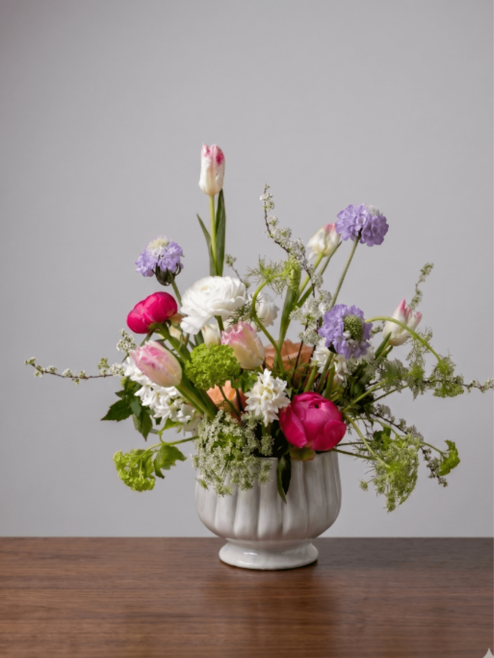 BP109 - Whimsical Spring Garden - This airy, garden-style arrangement gathers vibrant pink peonies, elegant tulips, and lavender scabiosa, perfectly accented by delicate touches of white spiraea and Queen Anne's lace in a classic white fluted bowl.  Seasonality: Peonies: Peak availability in late Spring through early Summer. Tulips &amp; Hyacinth: Classic early to mid-Spring blooms. Scabiosa: Late Spring through Summer. Spiraea &amp; Queen Anne's Lace: Spring through early Summer.  