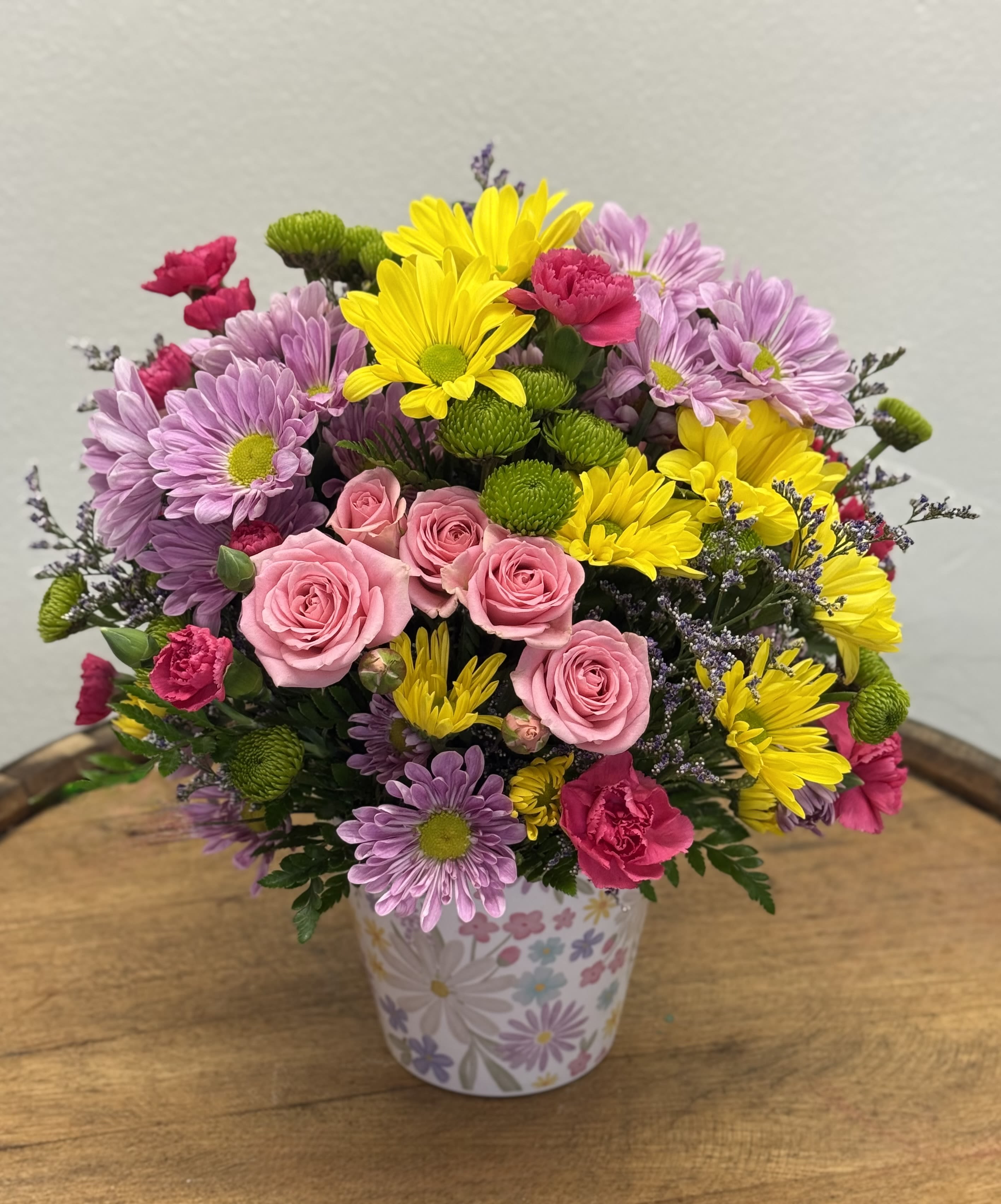 Sunshine Confetti - A burst of cheerful color-this flower arrangement pairs pink roses with bright yellow and lavender daisies, and hot pink carnations to create a lively birthday bouquet or thank-you gift that feels like a confetti celebration. The rounded, compact design reads joyful and intimate, the saturated pinks and sunny yellows balanced by soft purple tones so the recipient feels genuinely celebrated the moment they see it.  Arranged in a small floral-patterned decorative container rather than a formal vase, Sunshine Confetti has a casual, giftable charm perfect for desks, kitchen tables, or bedside stands. The mix of roses, daisies, and carnations gives a variety of textures-velvety rose petals, cheerful daisy centers, and ruffled carnations-packed into a neat, easy-to-place size.  Ideal for birthdays, thank-yous, or "thinking-of-you" moments, this compact arrangement delivers a joyful, uplifting impression without needing much space or fuss. Order with same-day delivery when available; keep the flowers in a cool spot away from direct sunlight and refresh the water (or replace the soaked floral foam) every couple of days to extend their bright, confetti-like presence.