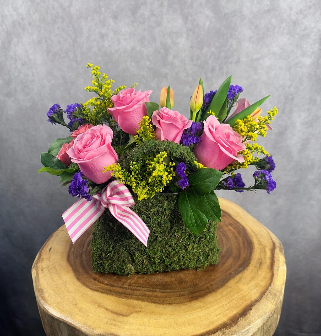 Pink roses and purple filler flowers arranged in a moss-covered box with a striped ribbon.