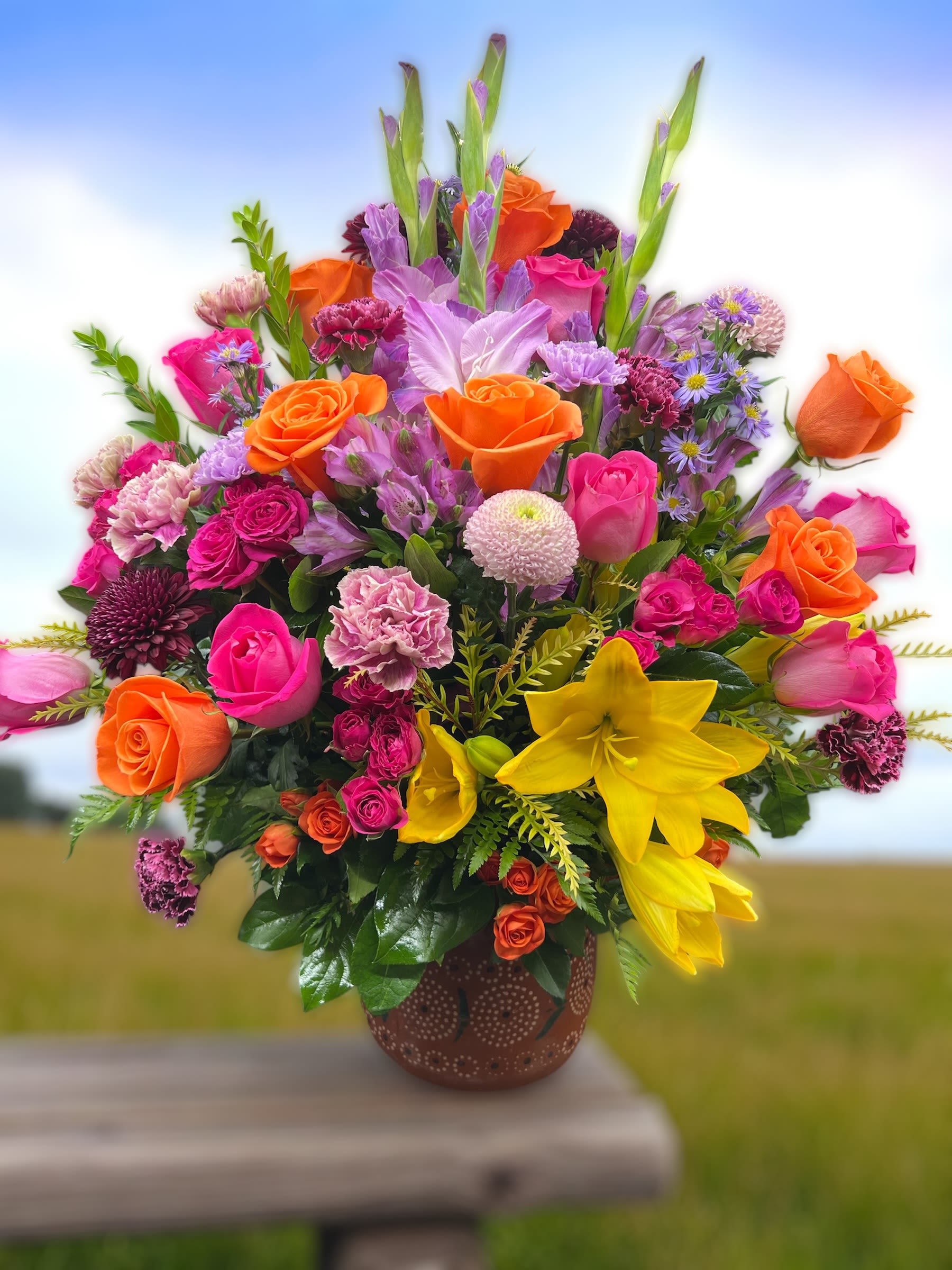 Tall mixed arrangement of pink and orange roses, yellow lilies, and purple accents in a brown pot