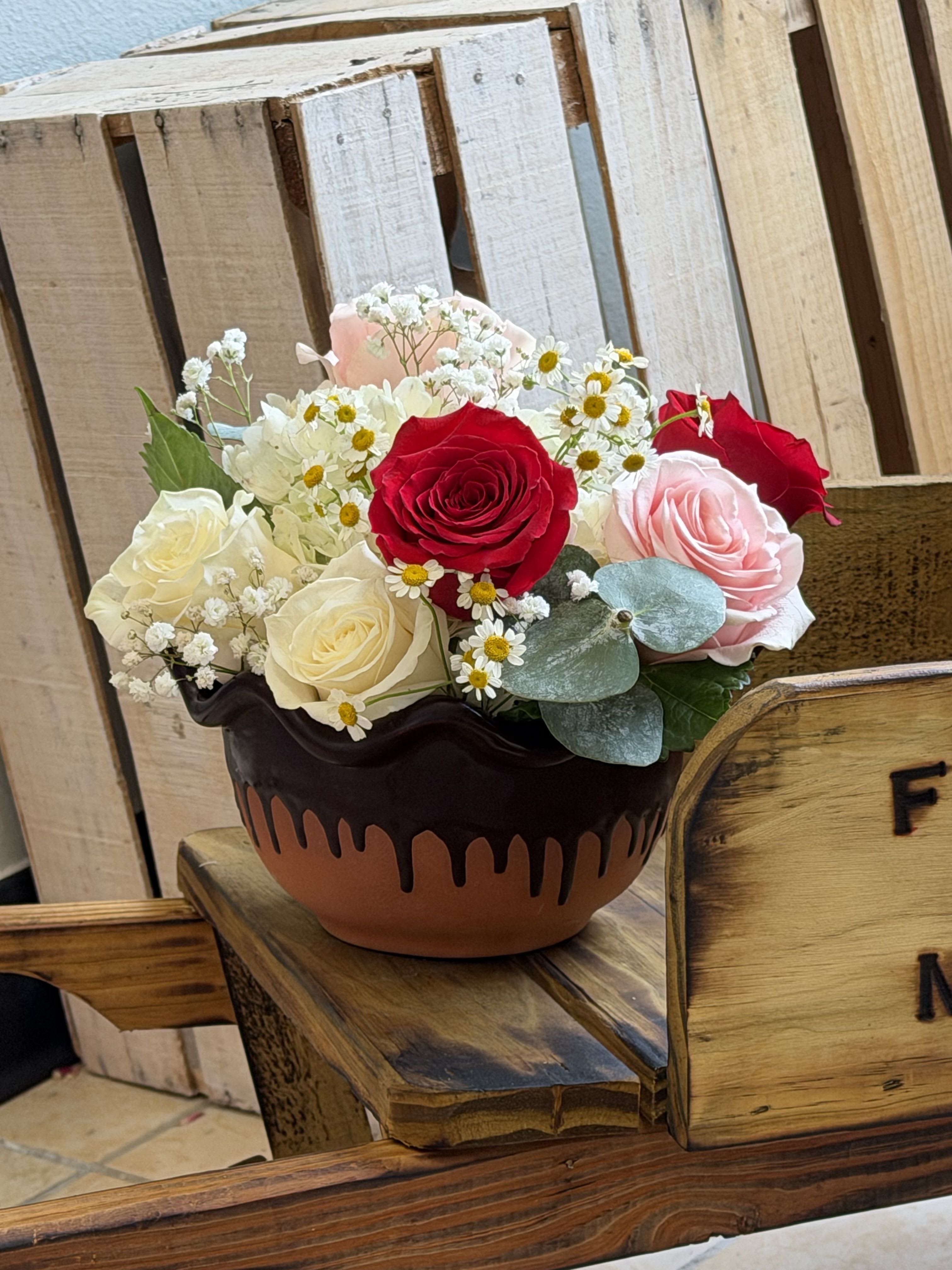 Delightful Dish - A compact cluster of red, blush pink and creamy white roses makes a romantic Mother's Day bouquet that reads both heartfelt and joyful. This Mother's Day arrangement is designed as a birthday-or-anniversary-worthy gift too-perfect for the mom who loves classic roses with a fresh, handmade feel-so the person you're celebrating will feel truly seen and cherished.  The roses are tightly gathered with small white daisy-like filler blooms and delicate baby's breath, softened by eucalyptus-like green foliage for cool contrast. Everything sits in a rounded ceramic bowl with a dark brown drip-glaze finish, giving the piece a rustic, handcrafted look. The composition is full and low-profile, ideal for a table centerpiece or keeps on a kitchen counter.  Give this to Mom as a meaningful way to say thank you and celebrate her day; the compact size makes it easy to place anywhere she'll see it often. Care is simple-keep water fresh and re-cut stems if possible; the ceramic bowl provides stable support and a long-lasting display of color and scent.
