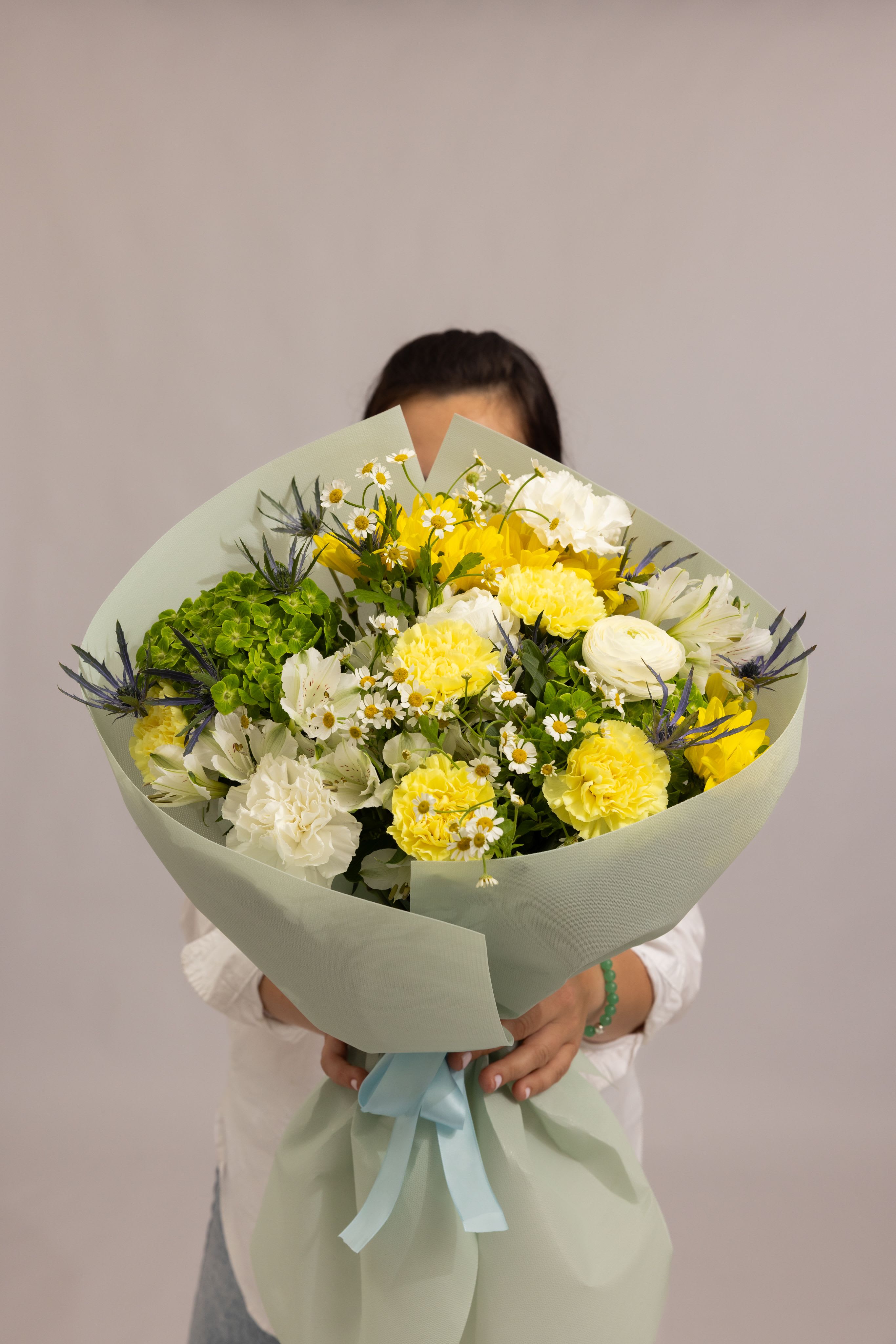 Lemon Whisper - A soft and refreshing bouquet in delicate yellow and white tones, featuring yellow and white carnations, white ranunculus, white alstroemeria, and daisy accents, complemented by a lush green hydrangea and subtle eryngium (blue thistle). The combination of textures and airy blooms creates a light, elegant composition that feels both modern and effortlessly graceful. Designed to bring freshness, warmth, and a gentle charm to any moment perfect for thoughtful gifts and refined celebrations.