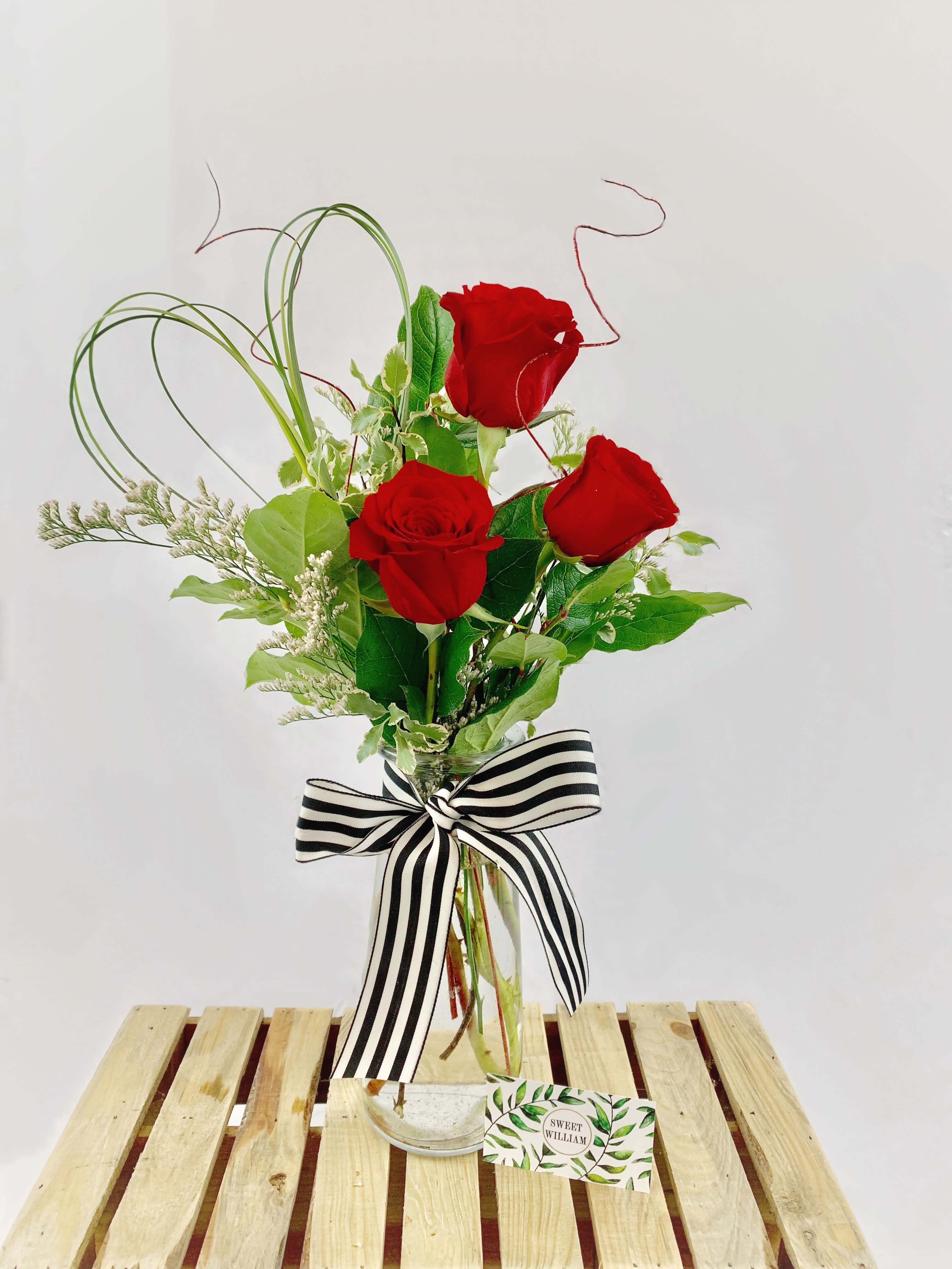 Three red roses in a clear glass vase with a striped bow