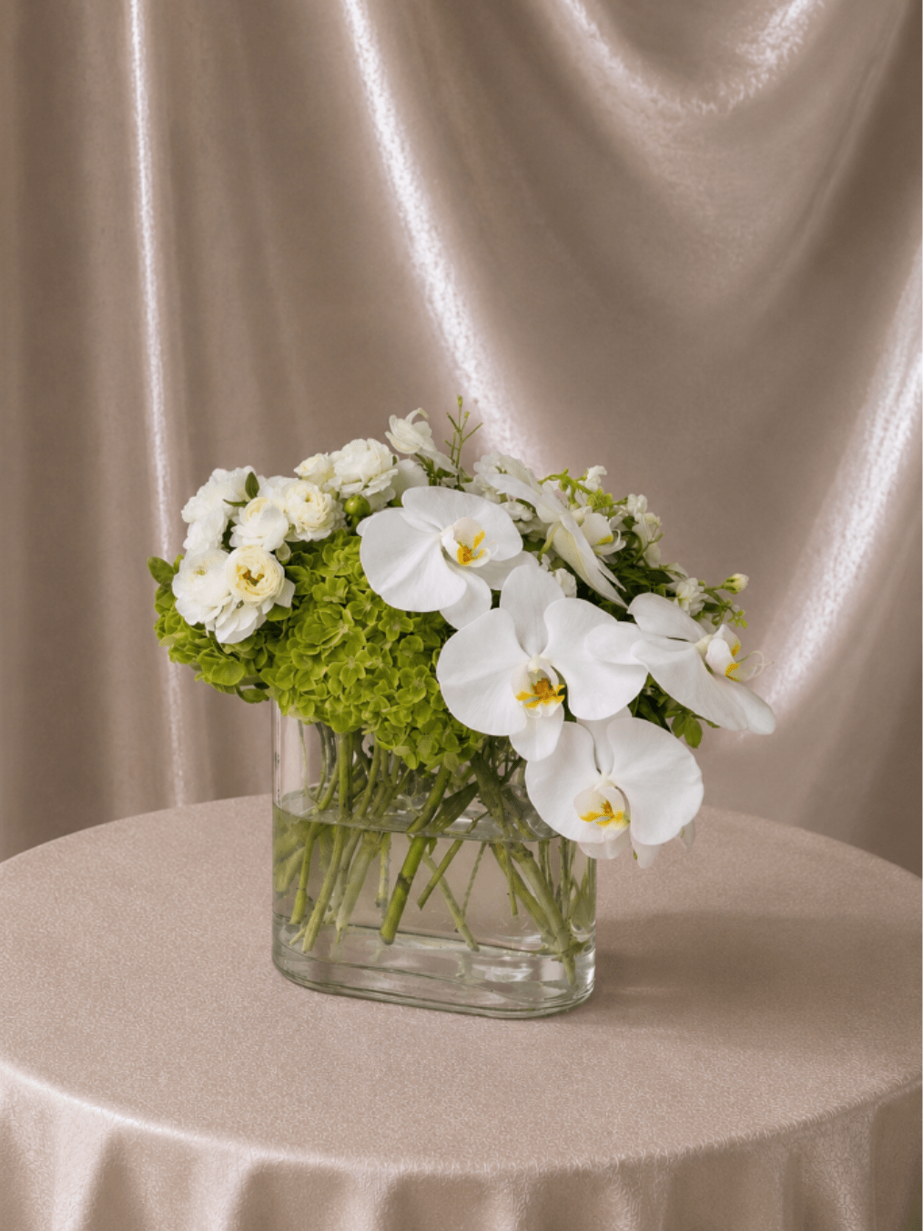 Elegant Orchid &amp; Hydrangea Garden - A refined and timeless flower arrangement featuring lush green hydrangeas, delicate white ranunculus, and premium white phalaenopsis orchids gracefully cascading over a clear glass vase. Blending softness and structure, this design creates a fresh, organic look with a touch of modern luxury.  Each arrangement is Designer’s Choice, thoughtfully created with the freshest seasonal blooms. Flower varieties and vase style may vary based on availability while preserving the overall look, palette, and refined composition.  Perfect for sympathy, get well wishes, thank you gestures, housewarmings, corporate gifting, birthdays, Mother’s Day, or adding a serene and elegant statement to any space.  Same day flower delivery available in Southwest Florida, including Naples, Bonita Springs, and Fort Myers Beach.