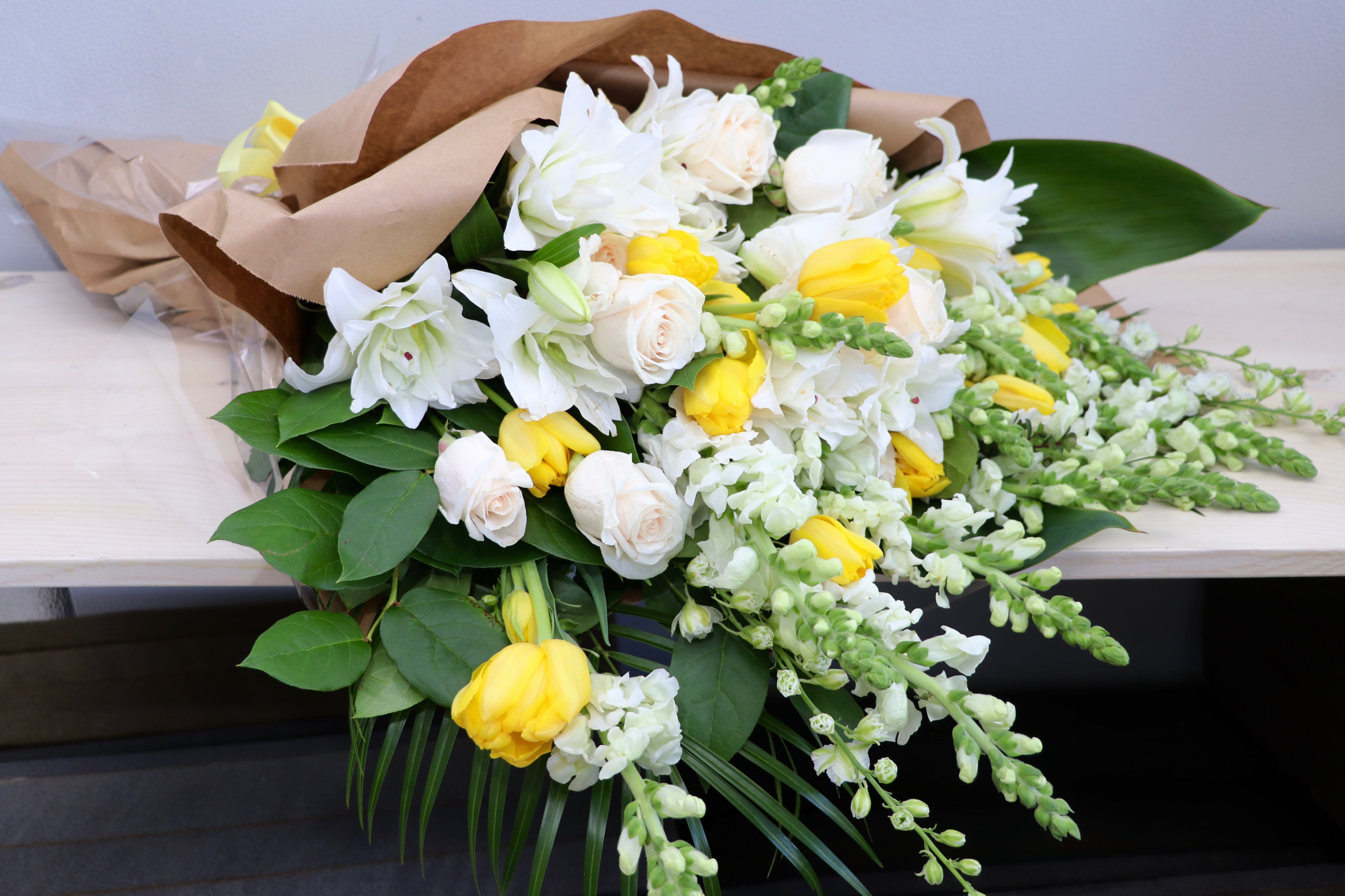 Wrapped bouquet of white lilies, cream roses, and yellow tulips with trailing white snapdragons