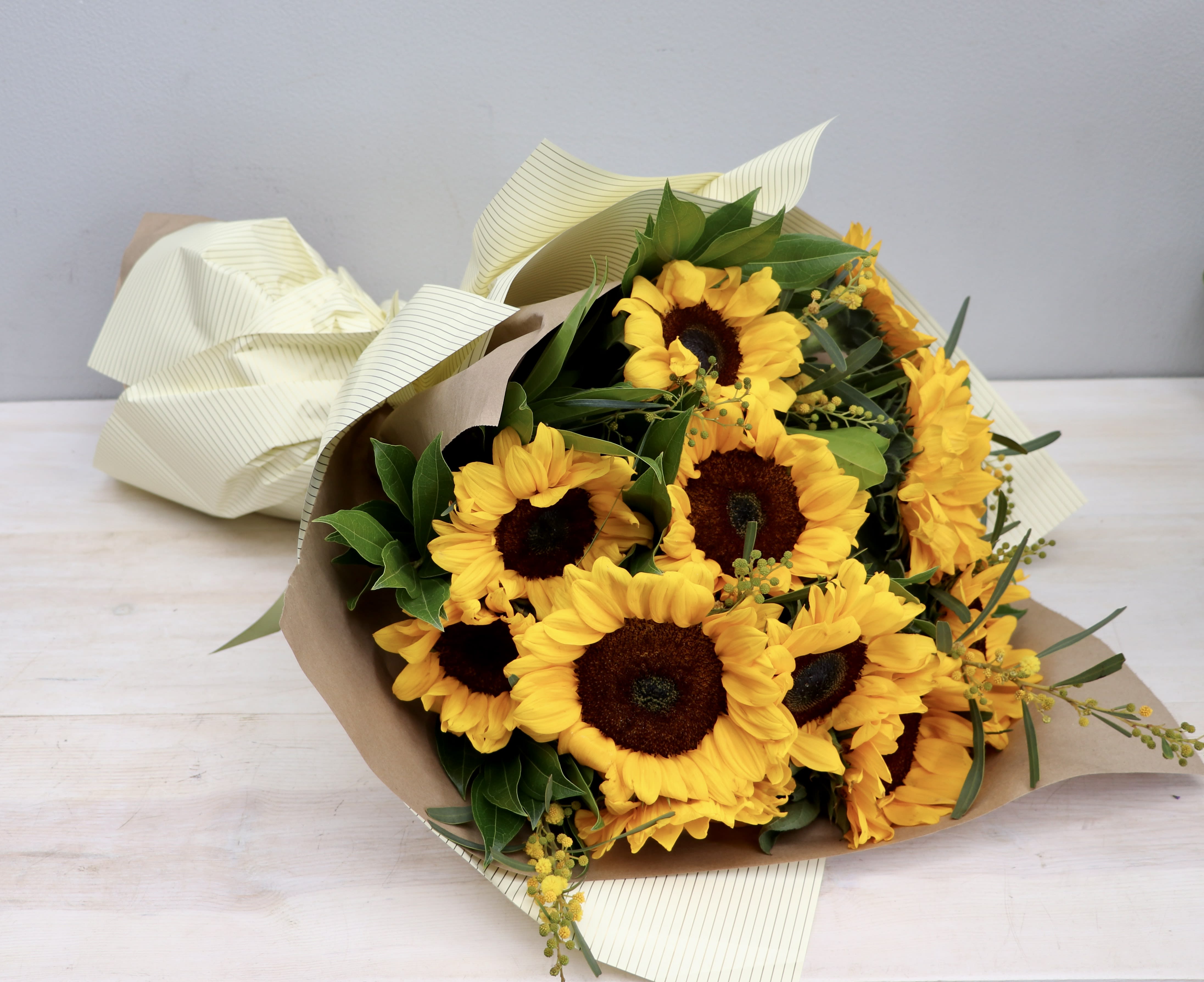 Wrapped bouquet of bright yellow sunflowers with brown centers on a light wood surface.