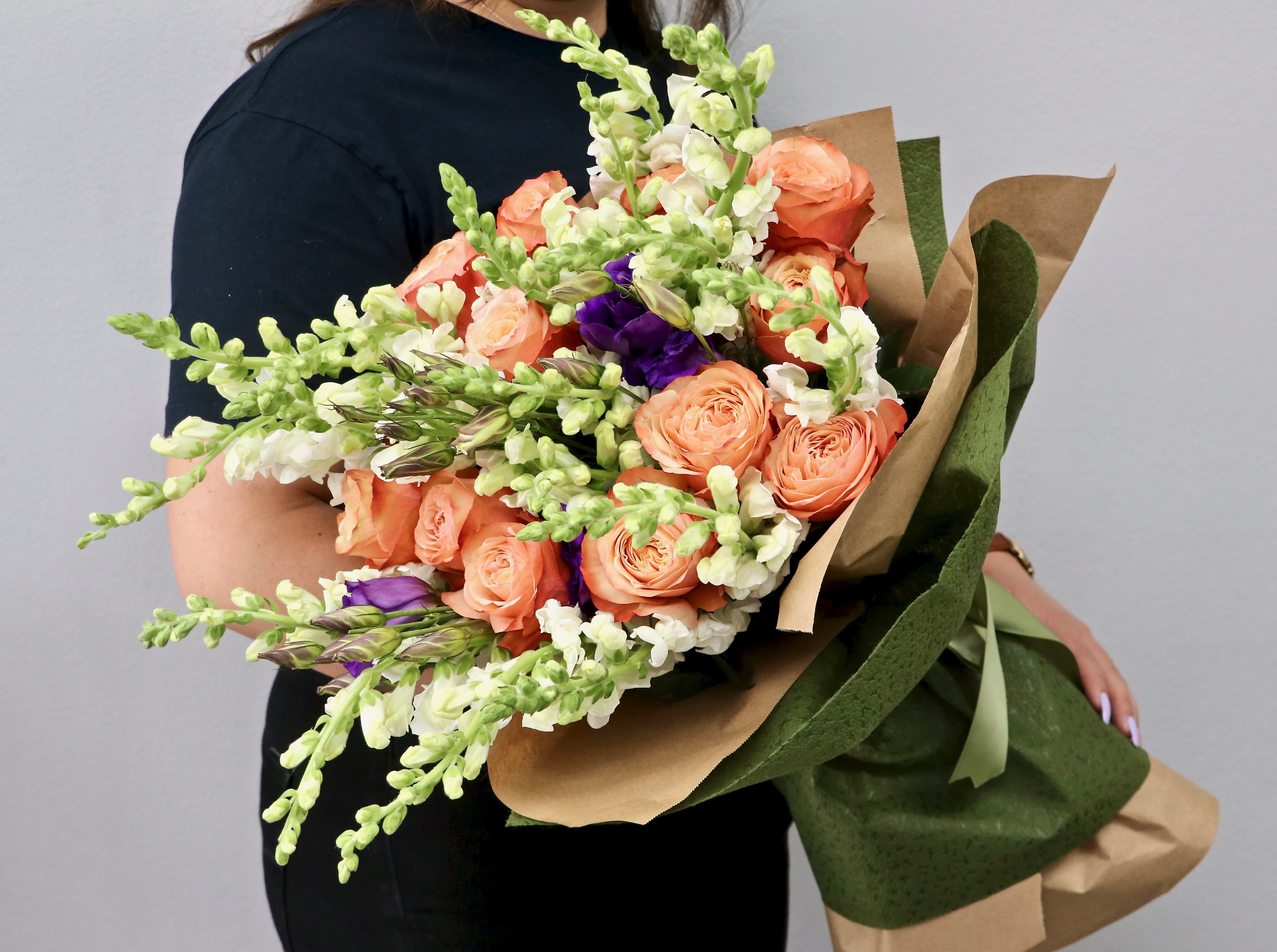 Large hand-tied bouquet of peach roses, white snapdragons, and purple flowers wrapped in kraft paper.
