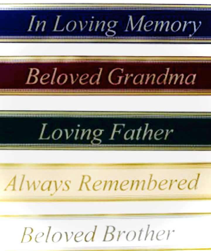 Banner Script  - Stacked memorial ribbon tags take center stage in this sympathy arrangement, offering a formal, respectful tribute for a funeral or Memorial Day remembrance. The Banner Script piece is designed to express condolence and honor - phrases like "In Loving Memory," "Beloved Grandma," and "Always Remembered" are printed on dark blue, burgundy, cream, and white ribbons with gold edging and serif lettering to create a solemn, dignified statement.  Beneath the ribbons, a full, rounded spray of white and pale yellow blooms - lilies, roses, and daisy-like chrysanthemums - forms a soft, serene backdrop. A wide black ribbon reading "Forever In Our Hearts" runs diagonally across the design, making the sentiment the focal point; the muted palette and classic flowers keep the arrangement appropriate for viewing and service settings.  Perfect for sympathy, funeral, or memorial services and meaningful for occasions like Grandparent's Day, retirement remembrances, or a heartfelt "I'm Sorry," this piece helps the person you're honoring feel truly remembered. Available as a sympathy add-on or standalone tribute; please note the prominent memorial ribbons and black accent make it most suitable for formal remembrance settings.