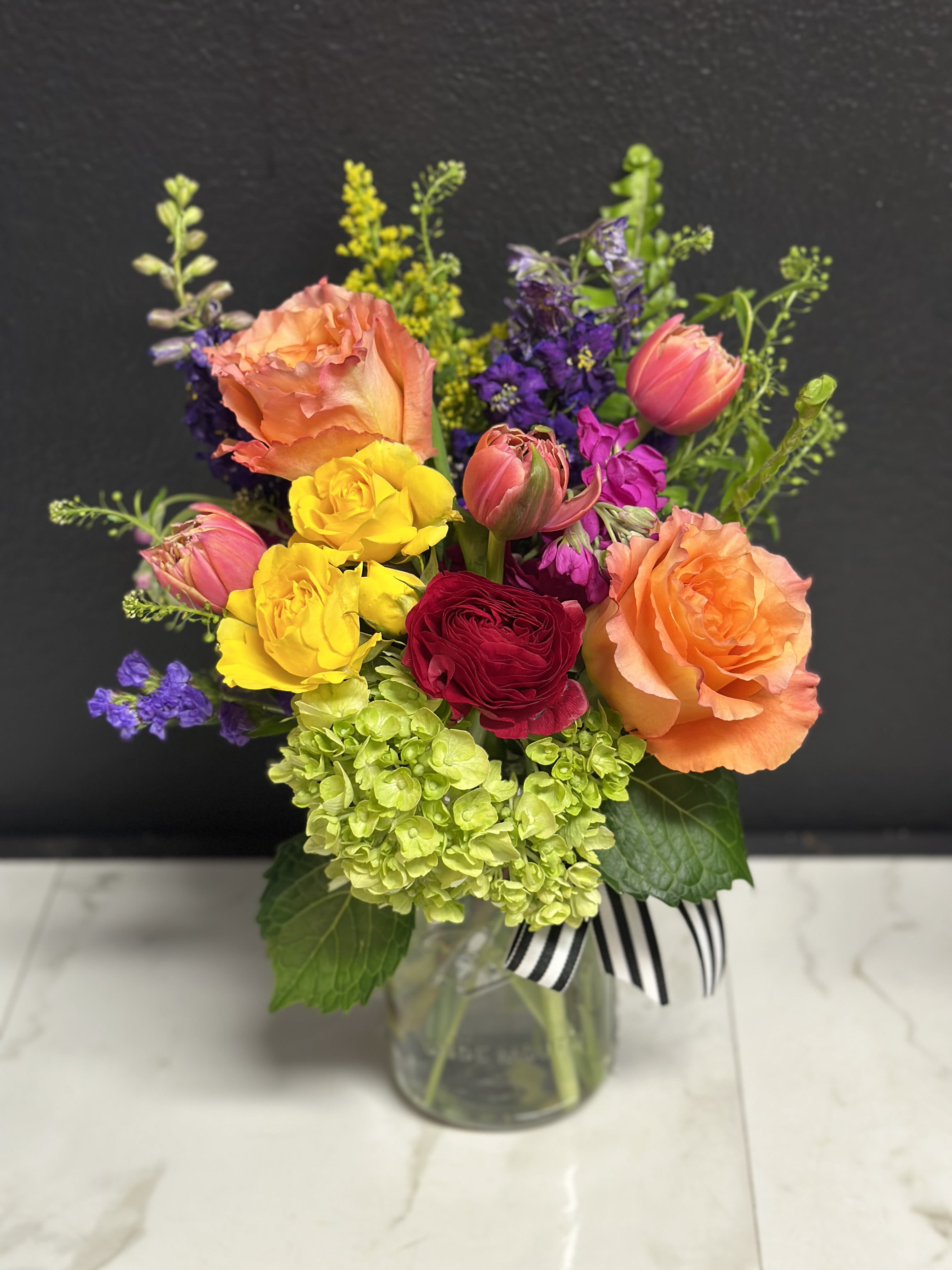 Vibrant Bloom Jar  - A bright, colorful, garden mix with season's prettiest blossoms. Arranged in our clear glass Bloom jar.              Overall design is approximately 12" tall.   Floral varieties may vary but will be of similar quality and style.  