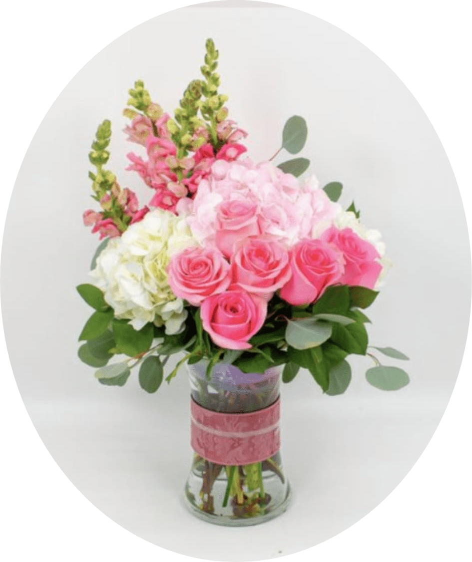 SIMONE - Soft blush roses and billowy hydrangeas meet tall pink snapdragon spikes in a romantic birthday bouquet that makes Mom, a friend, or a loved one feel truly celebrated. This flower arrangement for anniversaries or Mother's Day greets the room with soft pinks, creams, and fresh green accents-an instant expression of affection that reads as thoughtful and joyful.  The composition pairs pink and white roses with clusters of pale and white hydrangea, punctuated by vertical pink snapdragons for graceful height. Broad eucalyptus-like foliage adds texture and a fresh green contrast. Flowers arrive gathered in a clear glass vase wrapped with a pink decorative band and finished with a pink ribbon for a polished, gift-ready presentation.  Ideal for spring gifting, milestone moments, or simply saying "I'm thinking of you," this arrangement is designed to feel full and luxurious without being fussy. Vase dimensions and care instructions are standard: keep blooms in fresh water, trim stems occasionally, and refresh the water every two days to prolong the life of the roses, hydrangeas, and snapdragons.