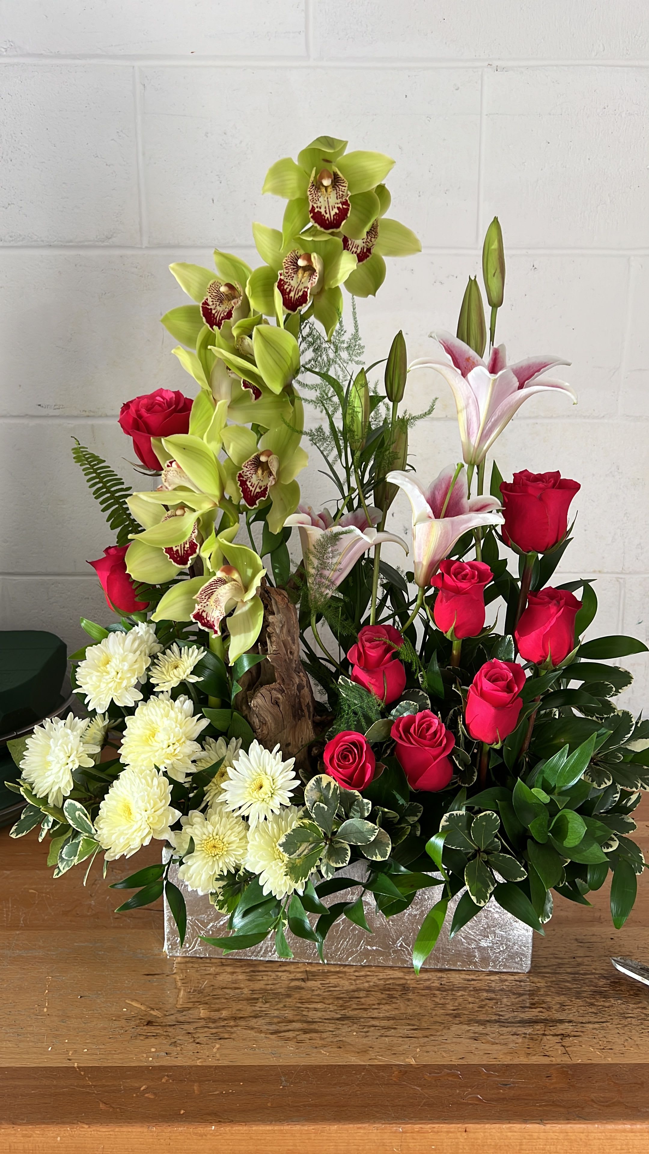 Mixed floral arrangement with red roses, green orchids, white lilies, and white mums in a silver box.