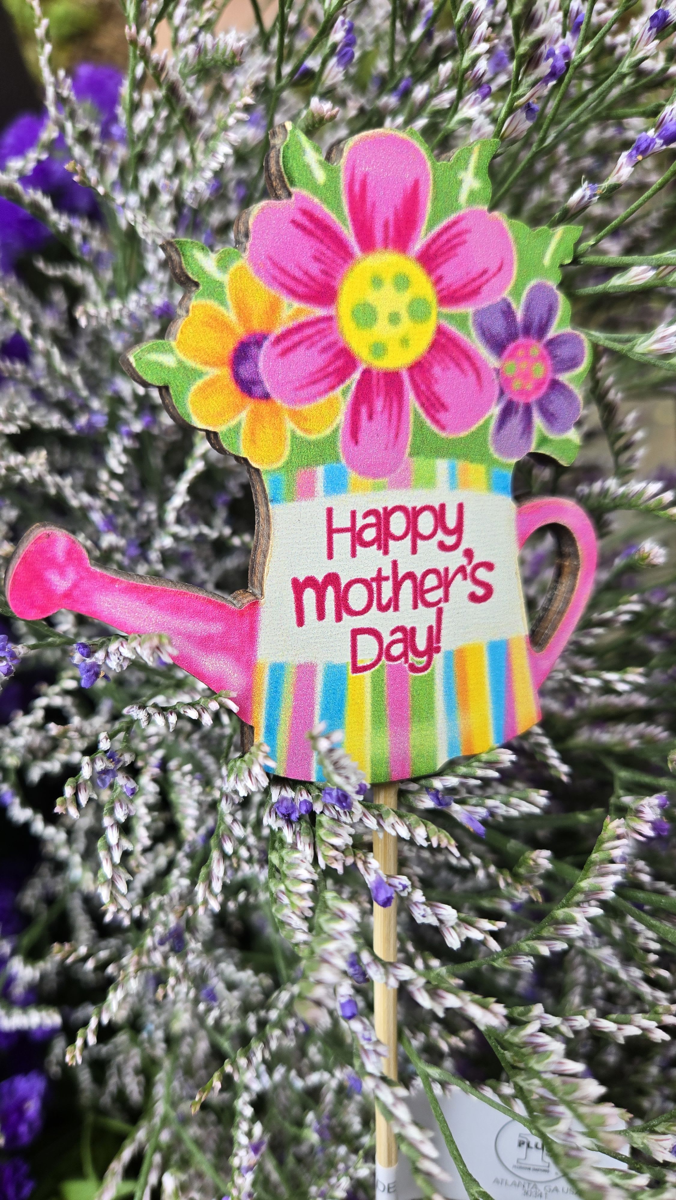 Happy Mom's  - A playful watering-can pick that reads "Happy Mother's Day!" crowns a dense mound of tiny purple-and-white blooms, making this Mother's Day flower arrangement feel bright, cheerful, and full of springtime charm. The vivid pinks and yellow accents on the wooden stem pick draw the eye and immediately announce the occasion, so the person you're celebrating will feel seen and celebrated the moment it arrives.  The arrangement features a textured mix of small purple and white filler flowers-soft, full clusters that give a lush, hand-gathered look-complemented by pops of green foliage and touches of yellow for contrast. A decorative wooden stem supports the colorful pick, adding a whimsical, keepsake element that continues to delight after the flowers are delivered.  Perfect for moms who love playful, seasonal arrangements or anyone you want to surprise with a sunny spring bouquet, this design is an easy, festive way to say "thank you" or "I love you" on Mother's Day. Order with confidence for delivery around the holiday; the pick and sturdy wooden stem help the arrangement retain its celebratory presentation through gifting.