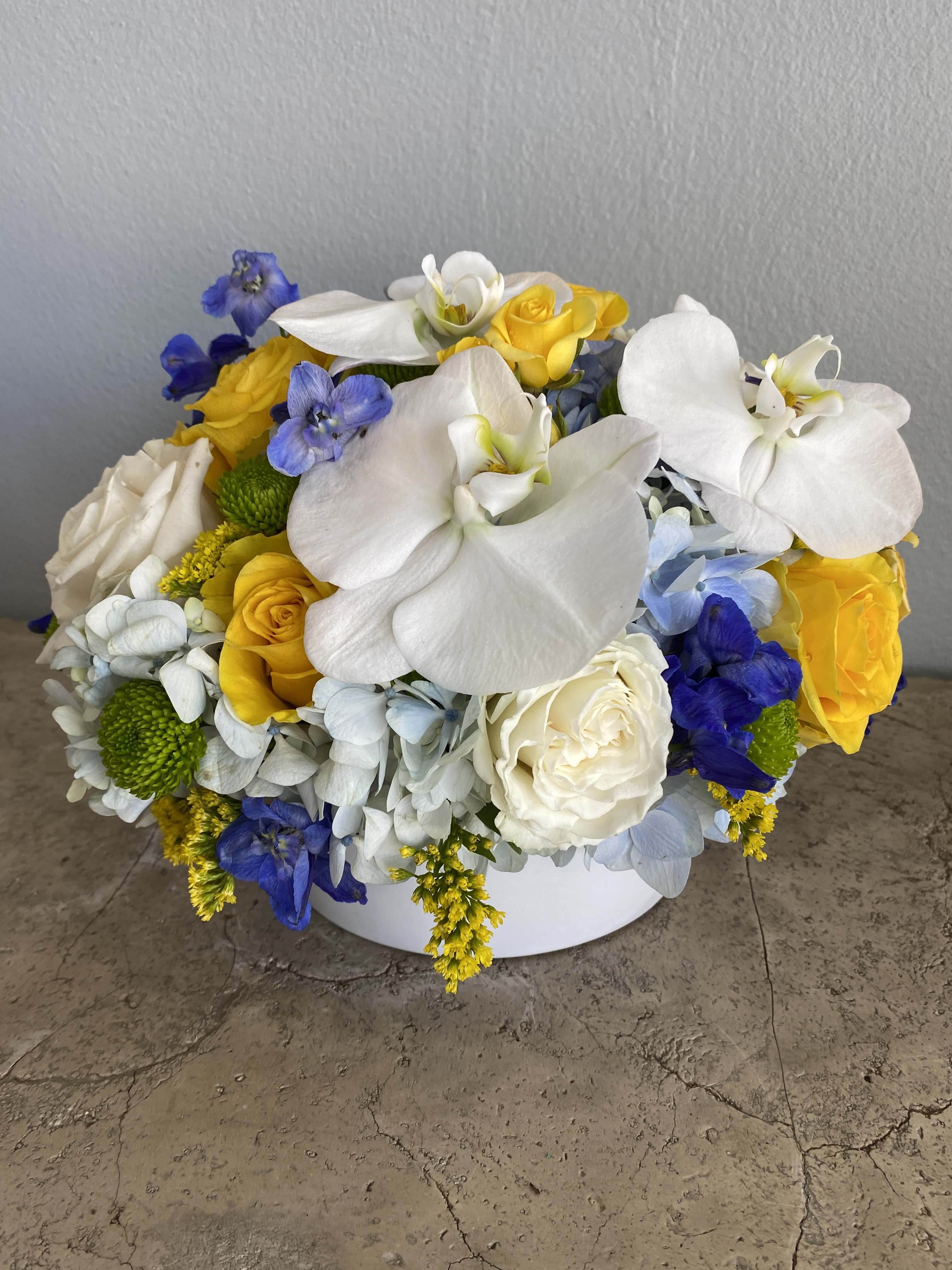 Round bouquet of white orchids, yellow roses, and blue flowers in a white container