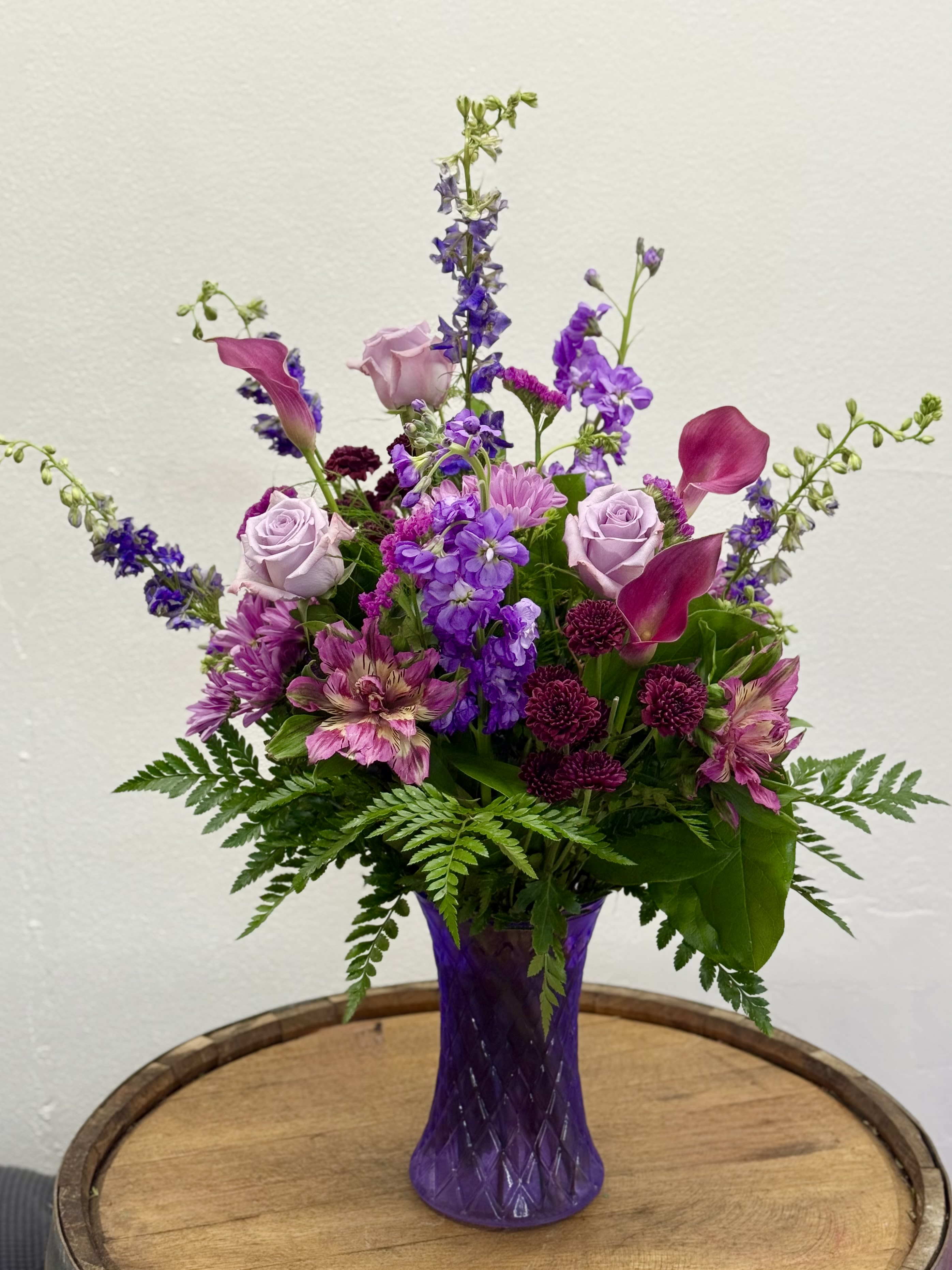 Amethyst Affection - Lavender roses and magenta calla lilies rise in a tall purple glass vase to create a lush, romantic flower arrangement perfect for an anniversary, birthday, or a thoughtful thank-you gift. The airy, vertical silhouette and the shades of purple give the recipient a sense of elegant surprise-they'll feel celebrated and truly seen when these jewel-toned blooms arrive.  This arrangement pairs soft lilac roses with vivid violet larkspur, lavender stock, pink/purple astronovas, and rich-hued calla lilies, accented by deep chrysanthemum touches and fern foliage that fans low around the base. The tall purple glass vase enhances the dramatic vertical lines and keeps stems supported while showing off its royal vibes; several stems extend above the main cluster for a graceful, garden-fresh look.  Ideal for someone who loves romantic color palettes or a bold everyday bouquet, it works equally well on a mantel, entry table, or dining centerpiece. Arrives arranged and ready to display; to keep blooms fresh, change the water every 2-3 days and trim stems at an angle when you refresh the vase.