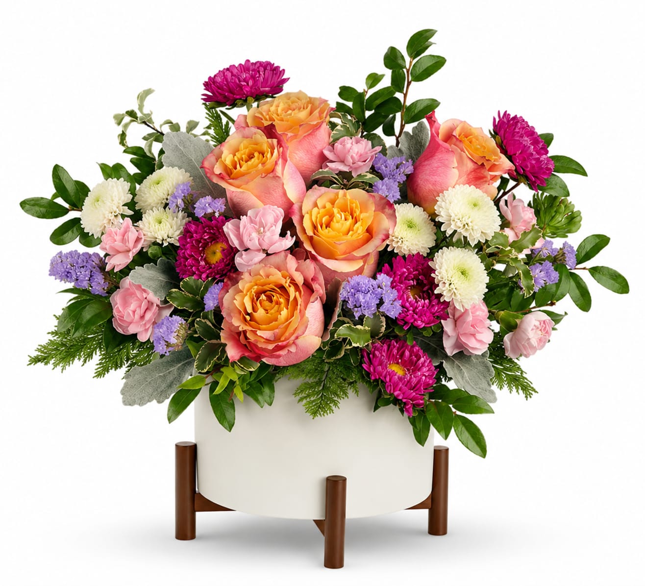 Blooming Iridescent Arrangement - Brighten someone's day with this charming floral arrangement, designed to bring warmth and joy to any occasion. Soft light orange roses take center stage, beautifully complemented by delicate miniature pink carnations. Hot pink Matsumoto asters add a vibrant touch, while crisp white button spray chrysanthemums provide contrast and freshness. Accents of lavender statice bring a hint of elegance, and lush floral greenery ties it all together.    Our pricing reflects both the flowers and the container chosen. Arrangements with premium vases or specialty materials may have a more compact floral design. If you’re looking for a fuller, larger look, we recommend choosing a Deluxe or Premium upgrade