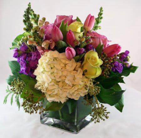 Mixed bouquet with white hydrangea, pink tulips, yellow roses, and purple flowers in a clear glass cube vase