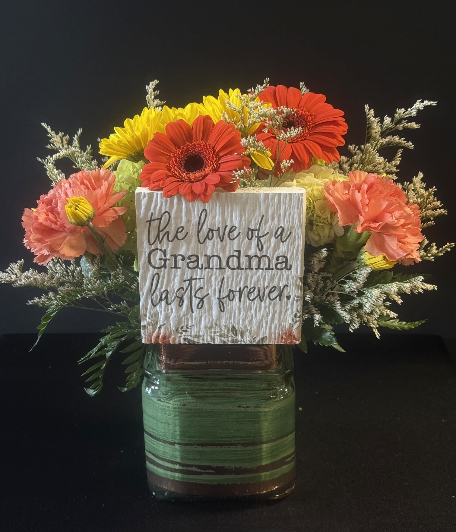 The Love Of A Grandma - Bright mixed gerbera, carnations and pale hydrangea in a glass vase with a rustic "the love of a Grandma lasts forever" sign-cheerful, heartfelt tribute in green, orange and yellows.