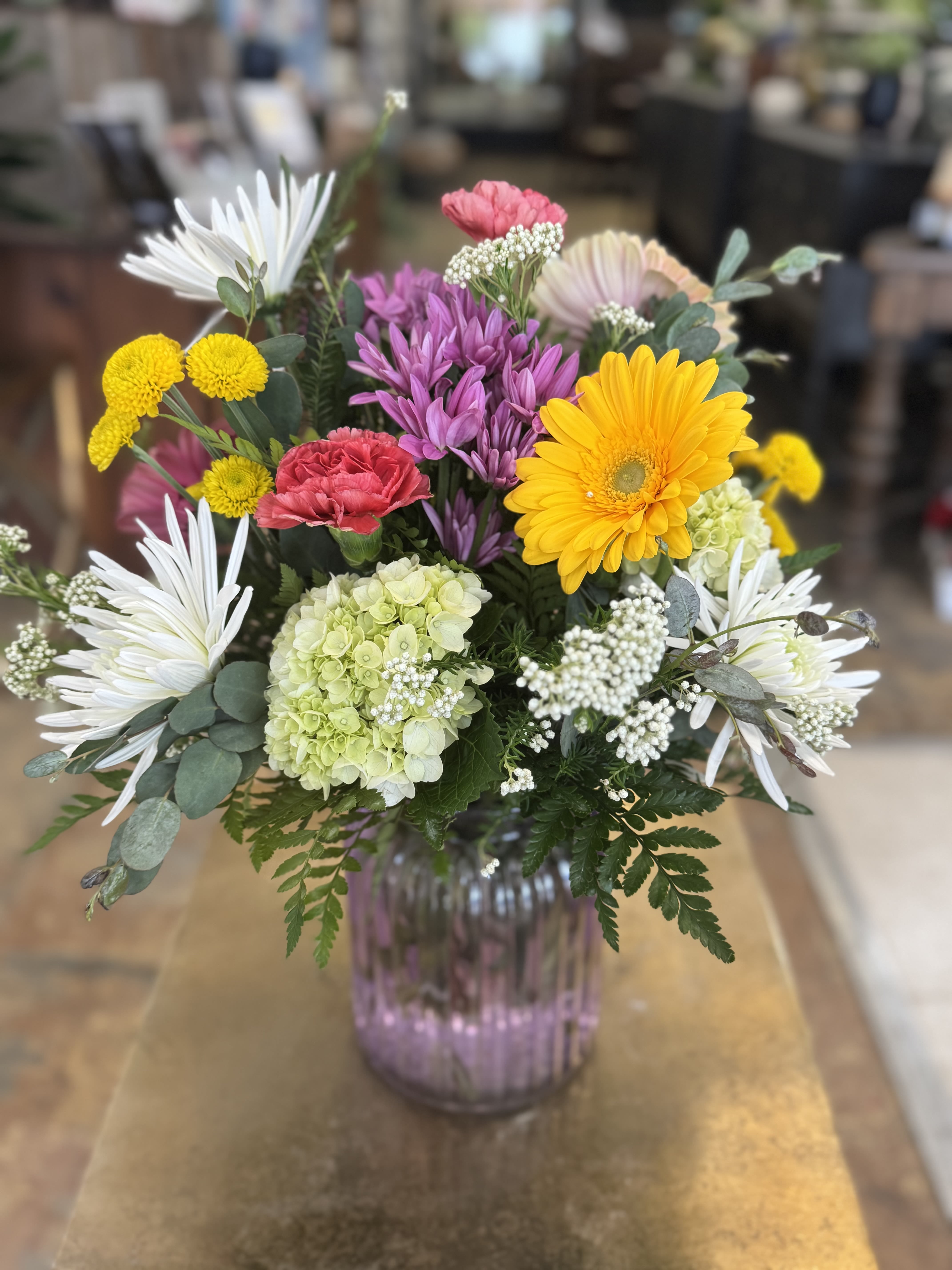 Mom's Garden Glow - A bright and beautiful Mother’s Day bouquet filled with cheerful yellow, pink, purple, and white blooms arranged in a soft lavender vase. Full of color, charm, and fresh garden-style beauty — a perfect way to celebrate Mom.