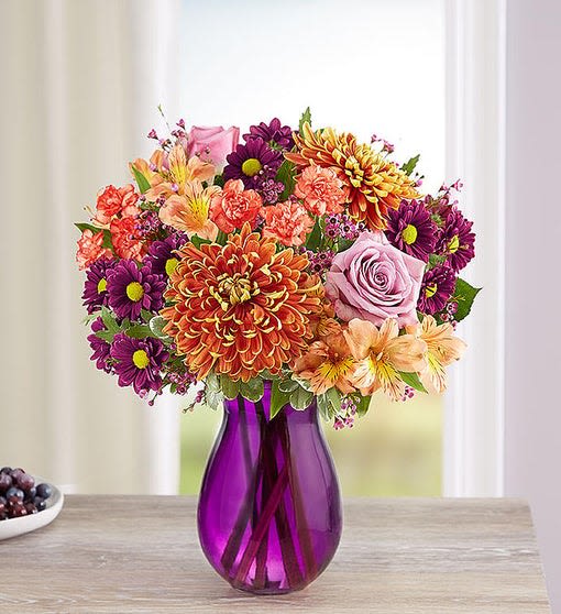 Mixed bouquet of purple, orange, pink, and peach flowers in a purple vase